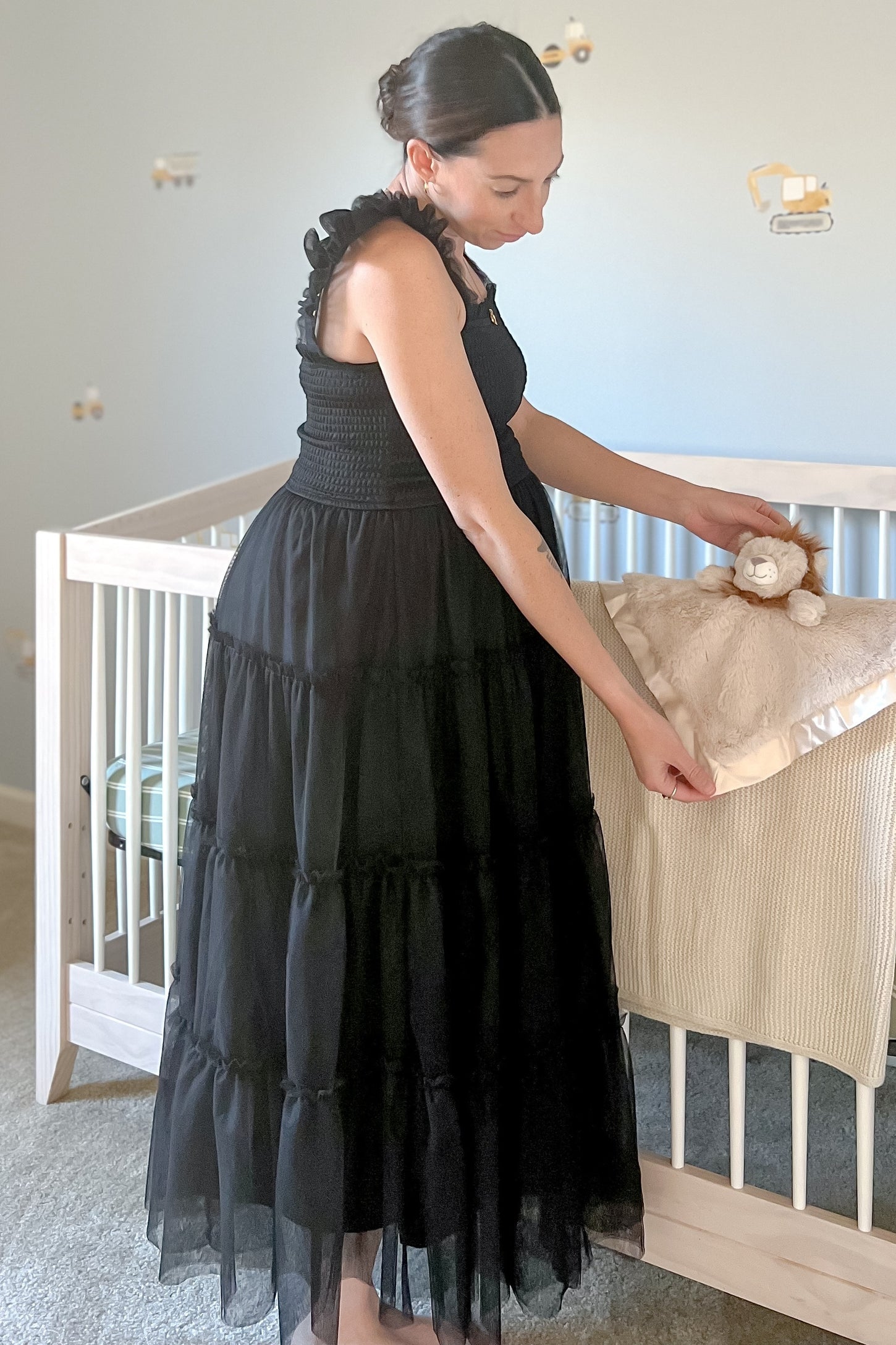 woman wearing a black tiered maternity maxi dress with ruffle sleeves, standing by a crib and holding a plush toy