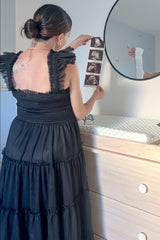 woman wearing a black maternity dress with ruffled straps, holding ultrasound photos in front of a mirror