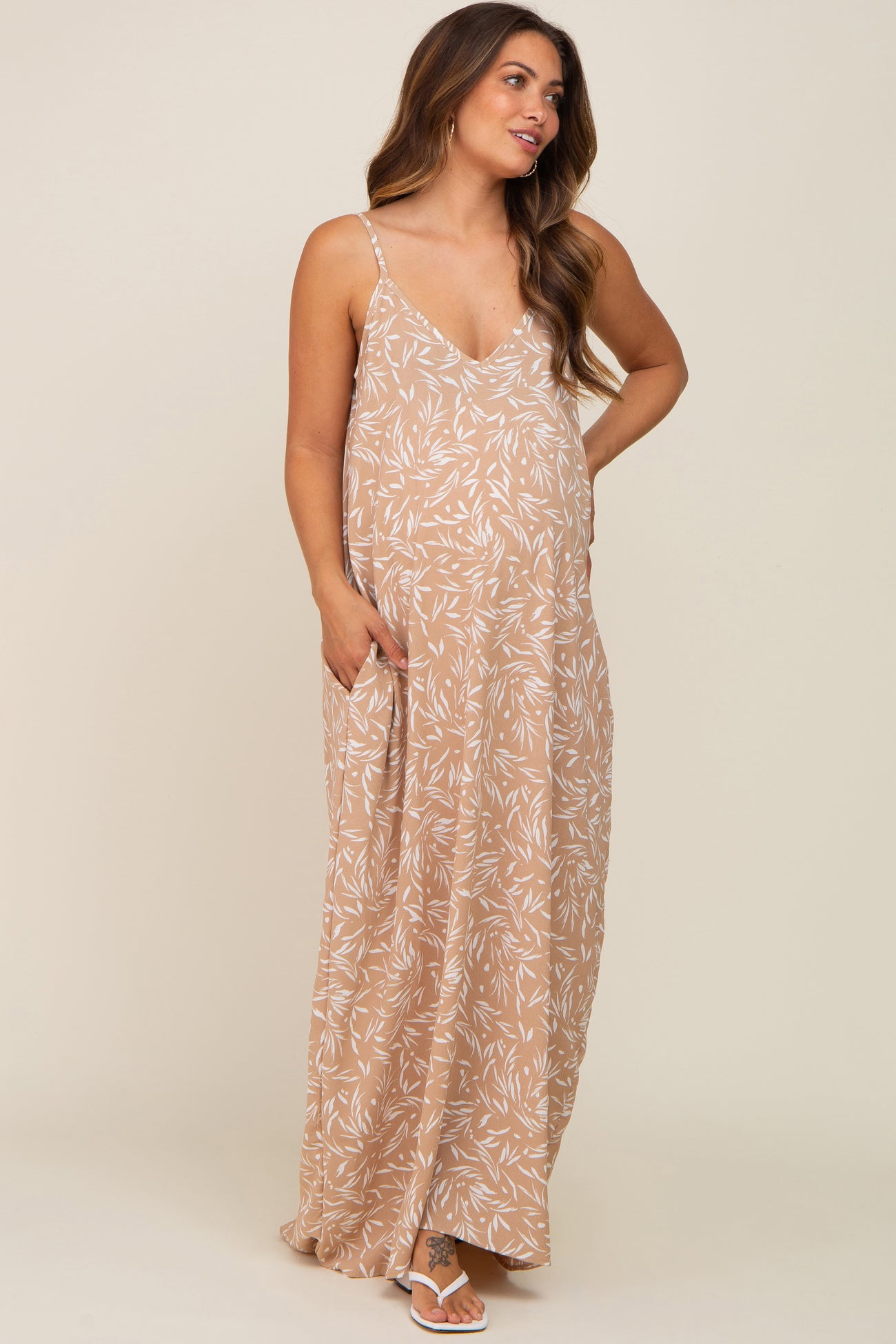 woman wearing a tan floral maternity maxi dress with adjustable straps and side pockets, showcasing her baby bump