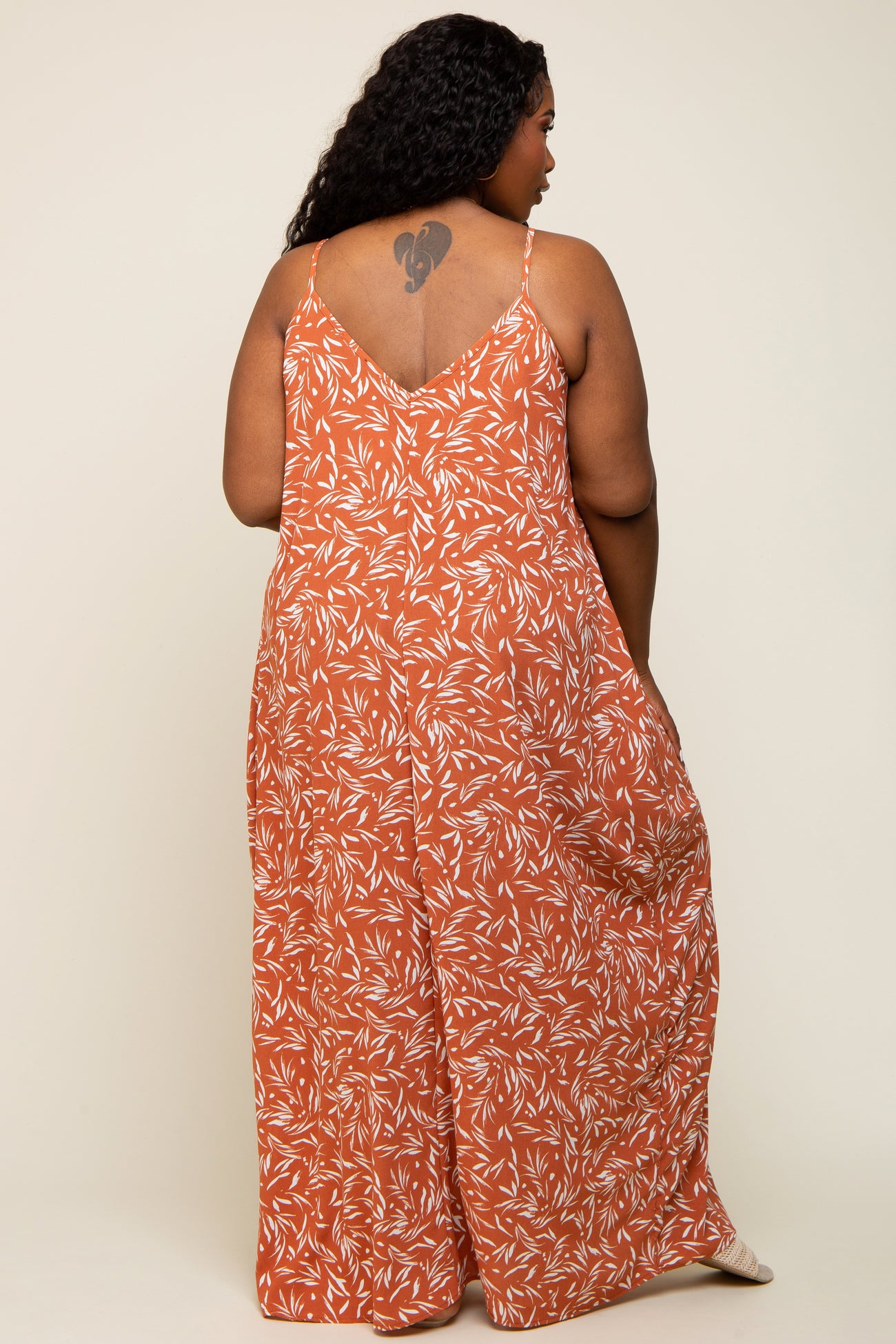 woman wearing an orange floral patterned plus-size jumpsuit with adjustable spaghetti straps, showing a tattoo on her back