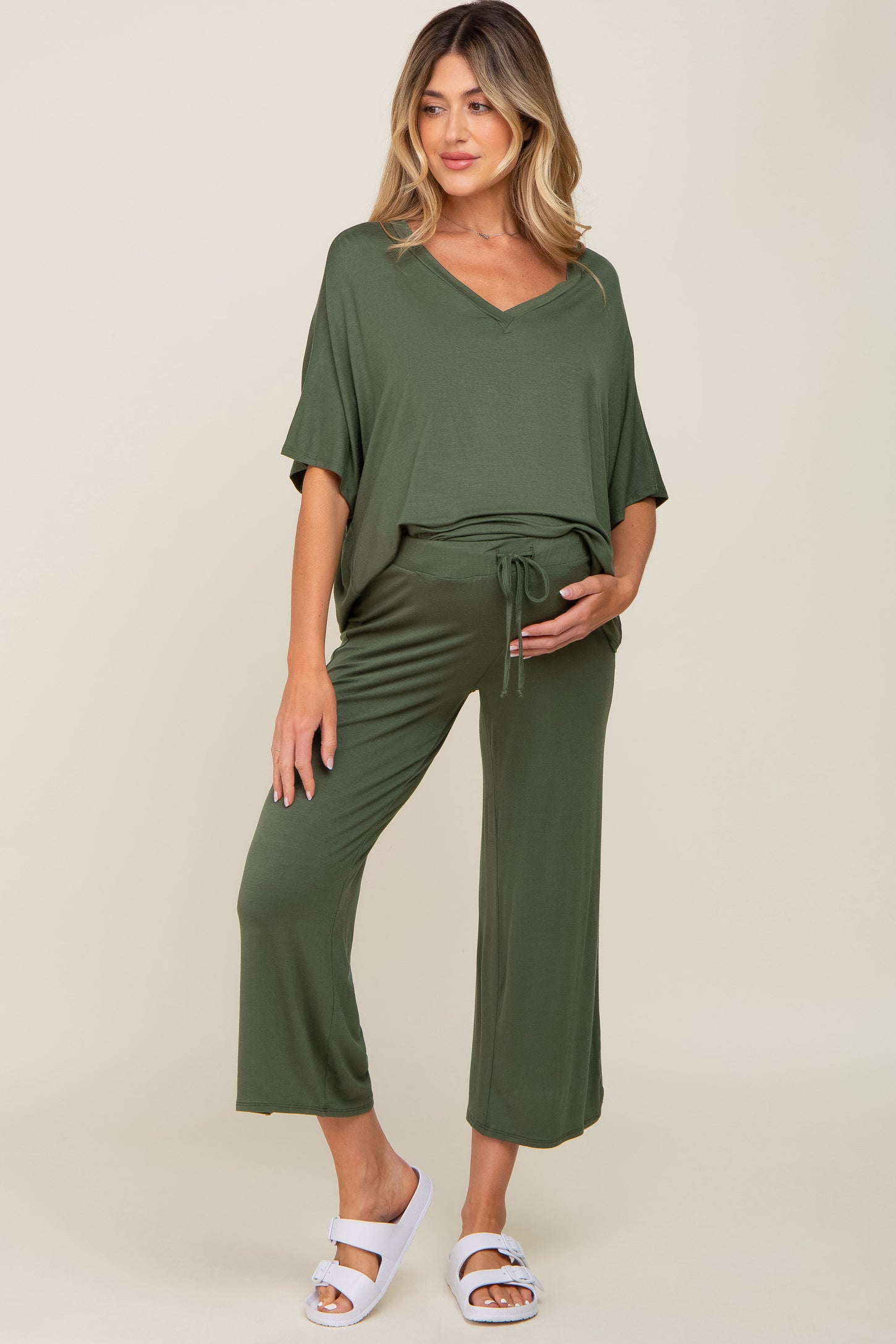 woman wearing an olive green maternity top and matching pants, showing her baby bump while standing on a neutral background
