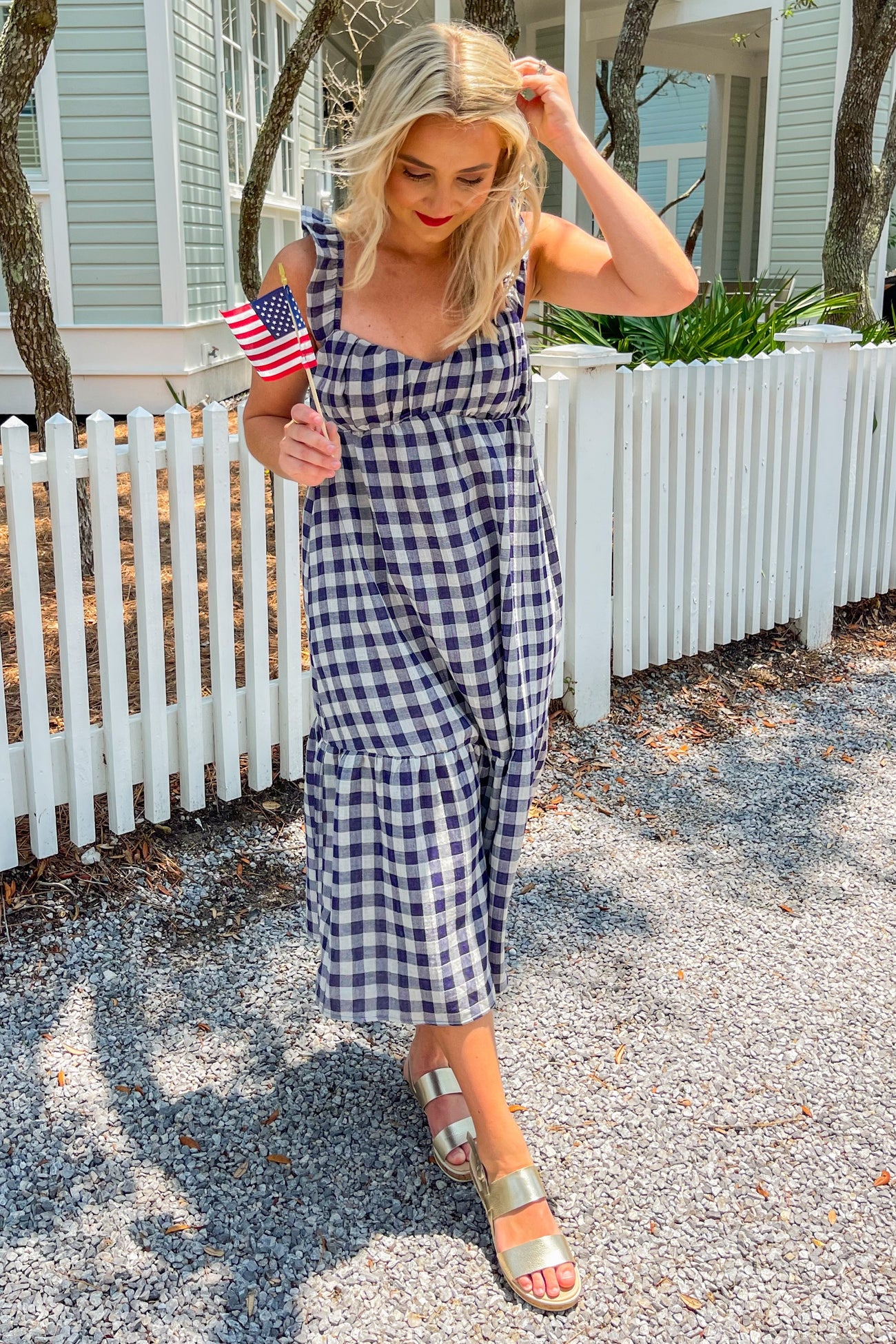 woman in navy and white gingham midi dress holding a small american flag by a white picket fence