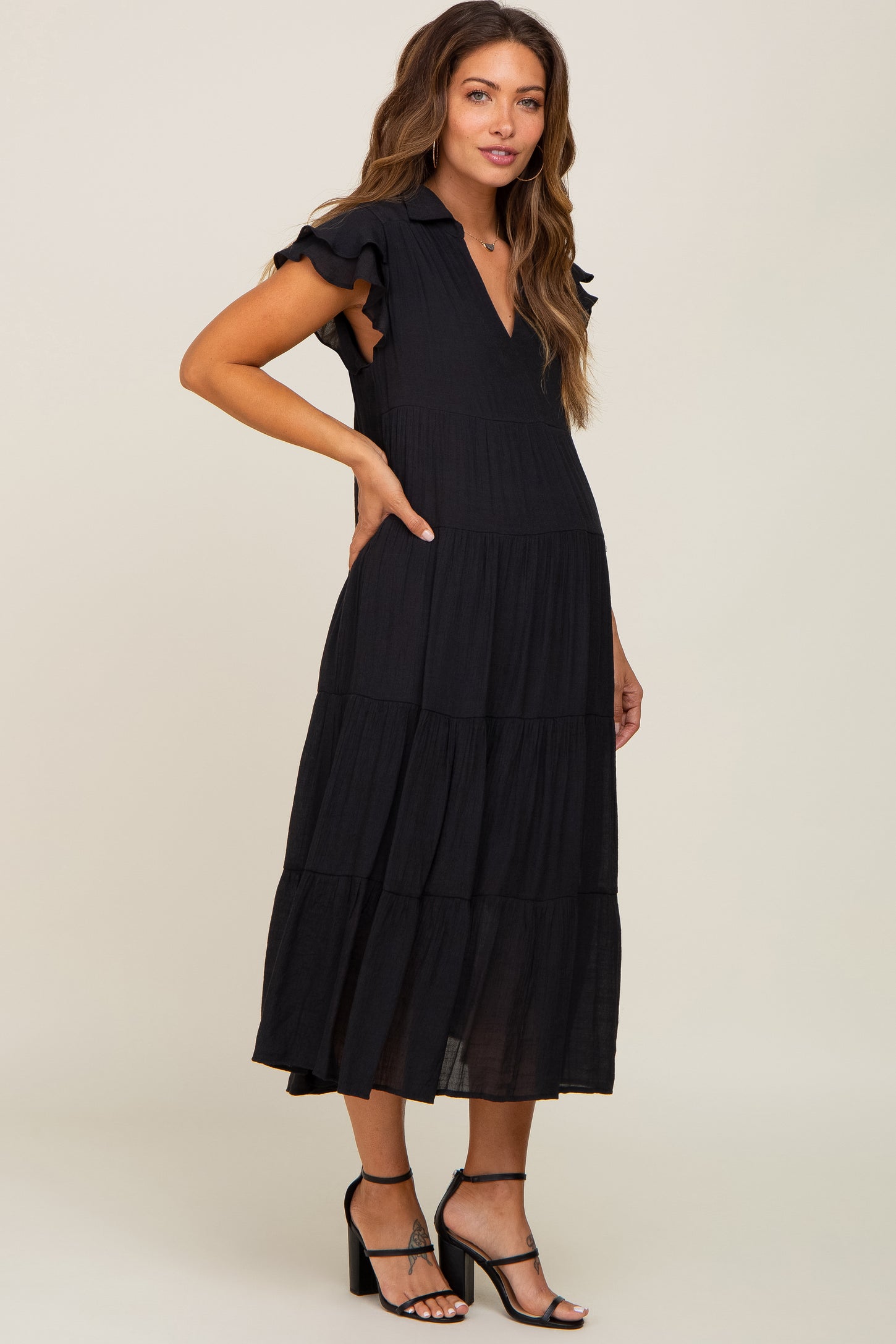 woman wearing a black tiered maternity midi dress with ruffle sleeves and black strappy heels
