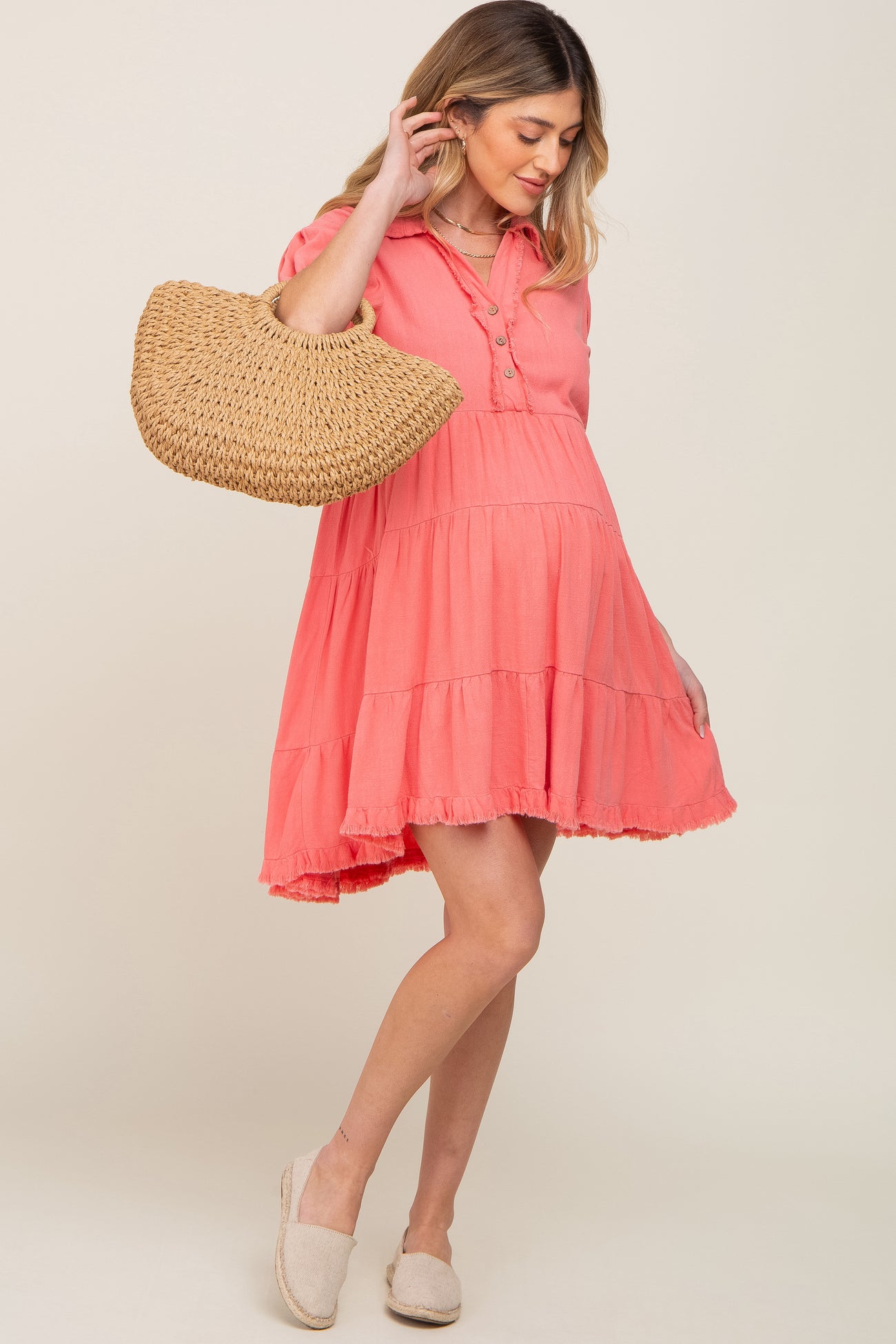 woman wearing a coral maternity dress with tiered design and holding a straw bag, baby bump visible