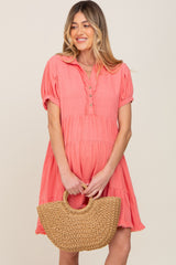 woman wearing a coral pink maternity dress with short sleeves and a tiered design, holding a straw bag and showing a baby bump