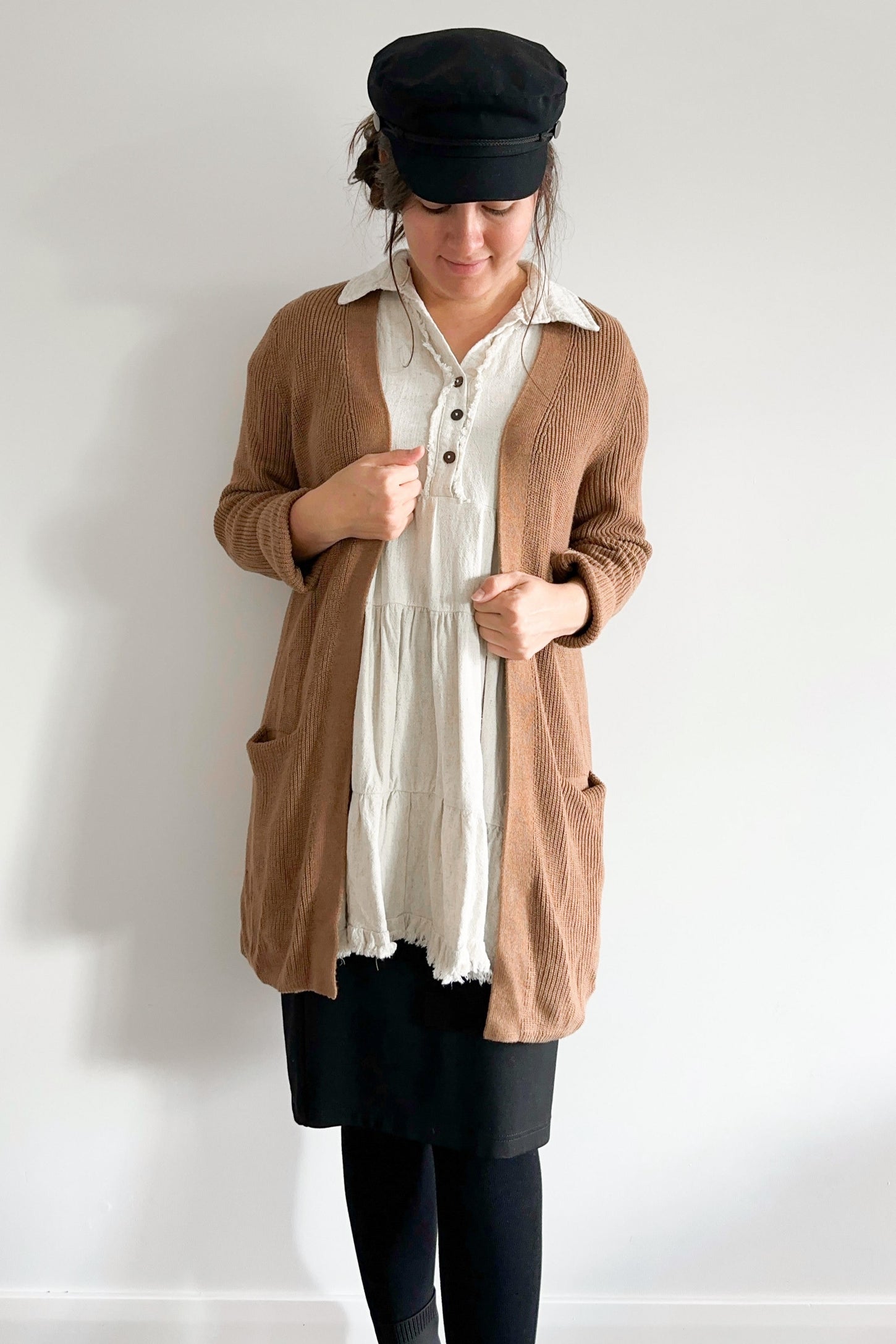 woman wearing a brown knit cardigan over a white tiered tunic dress and black baker boy hat