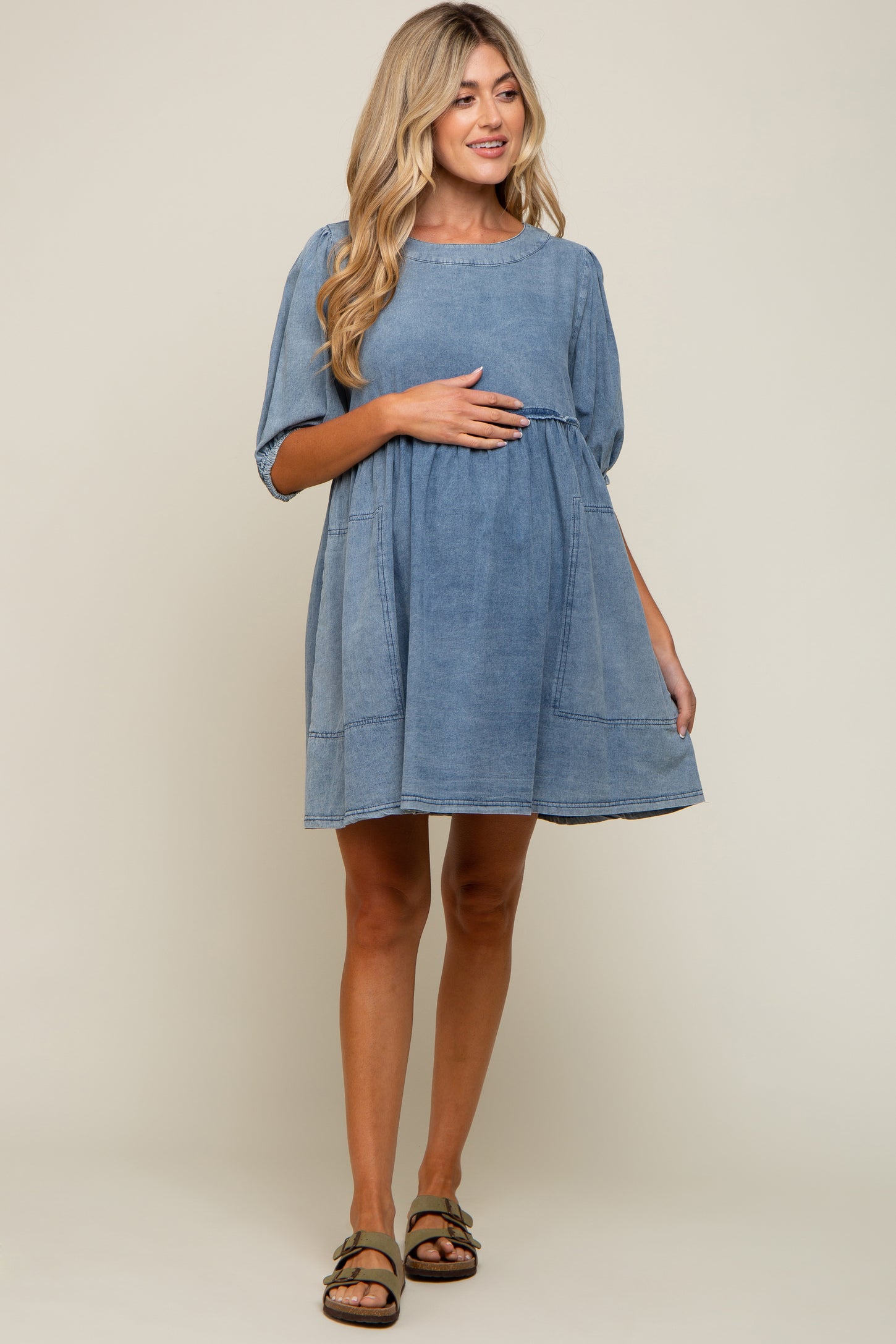 woman wearing a light blue denim maternity dress with short sleeves and a gathered waist, holding her baby bump