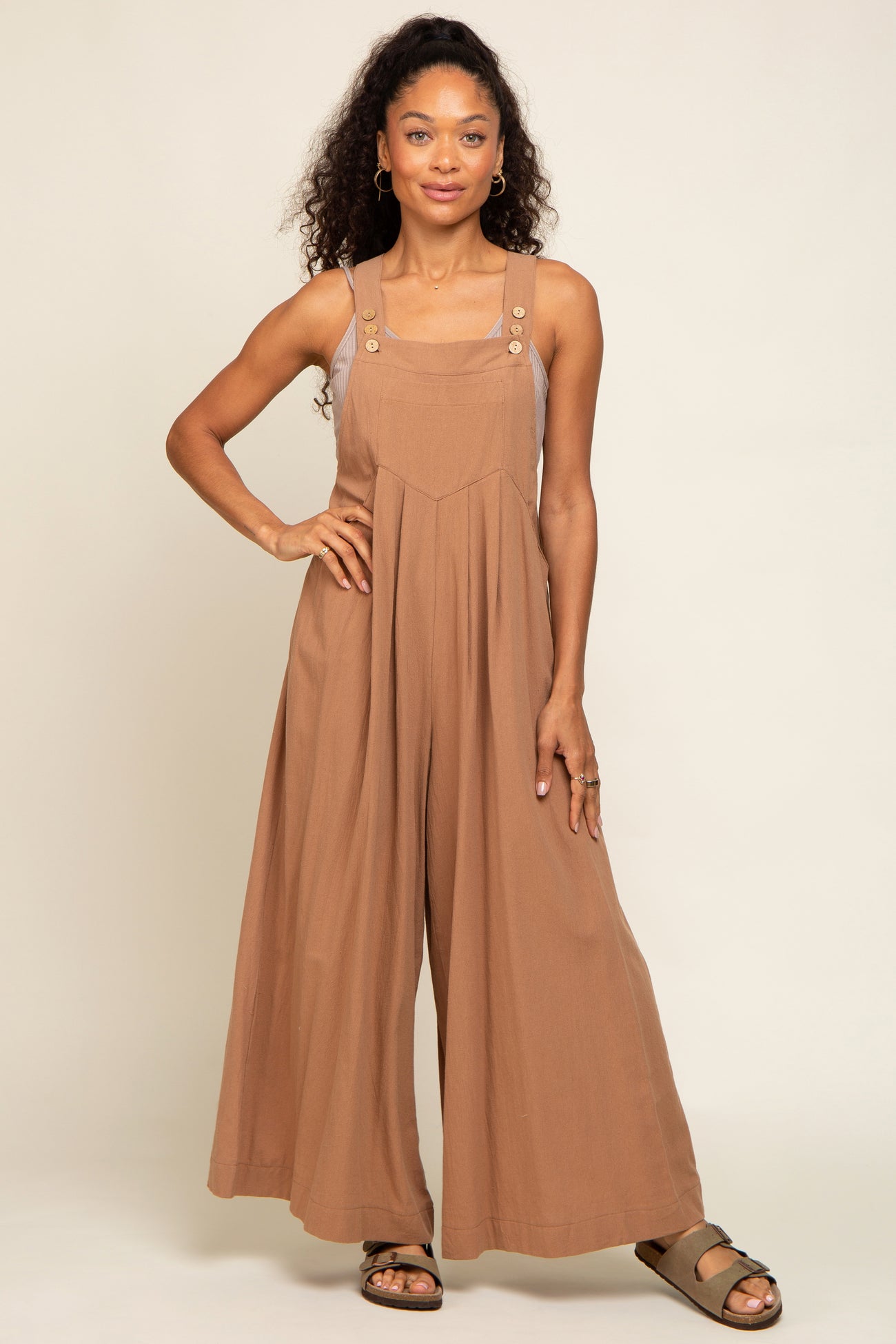Woman wearing a brown wide-leg jumpsuit with button straps and pleated front against a neutral background
