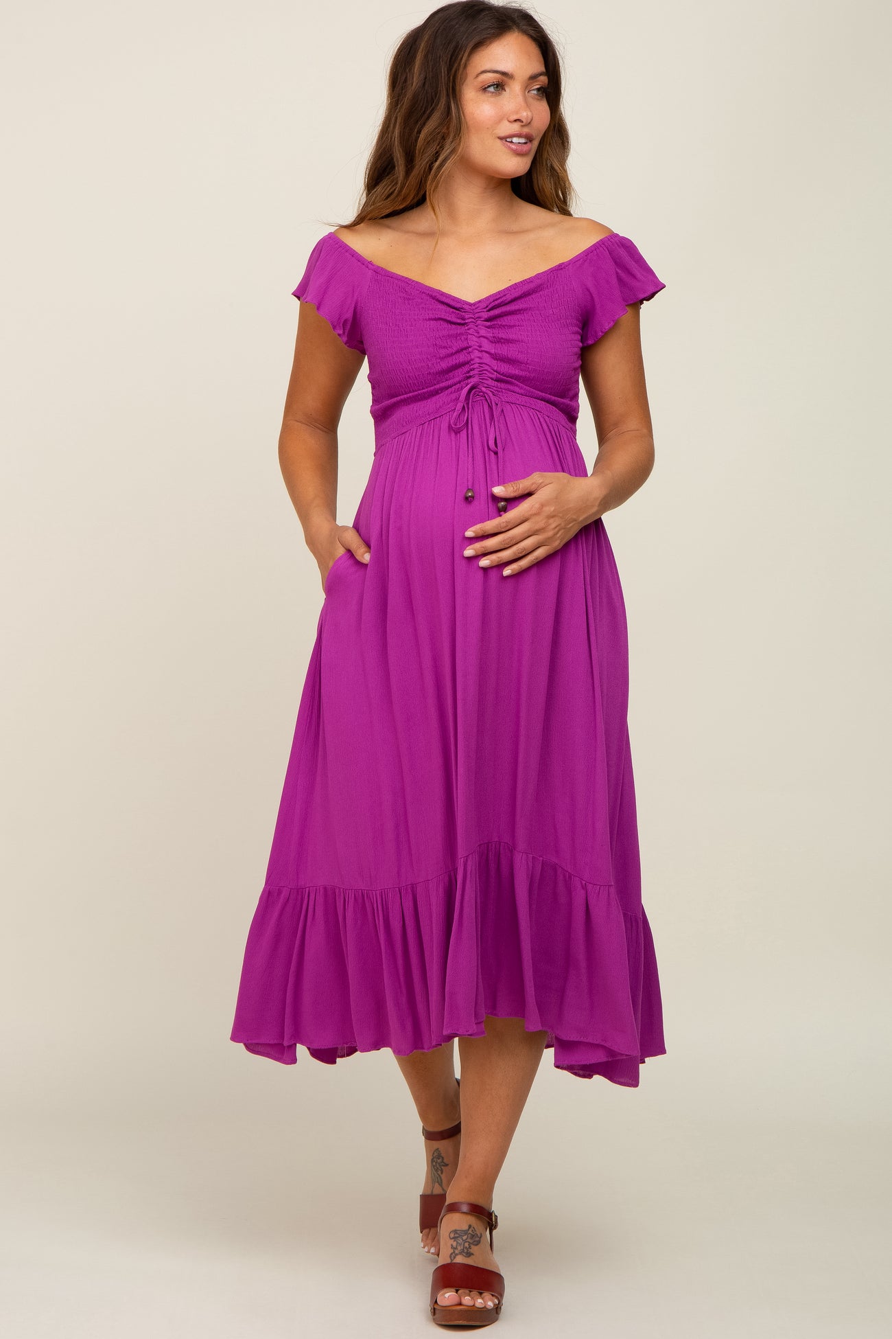 woman wearing a fuchsia off-the-shoulder maternity dress with a tie-front detail and ruffled hem, holding her baby bump