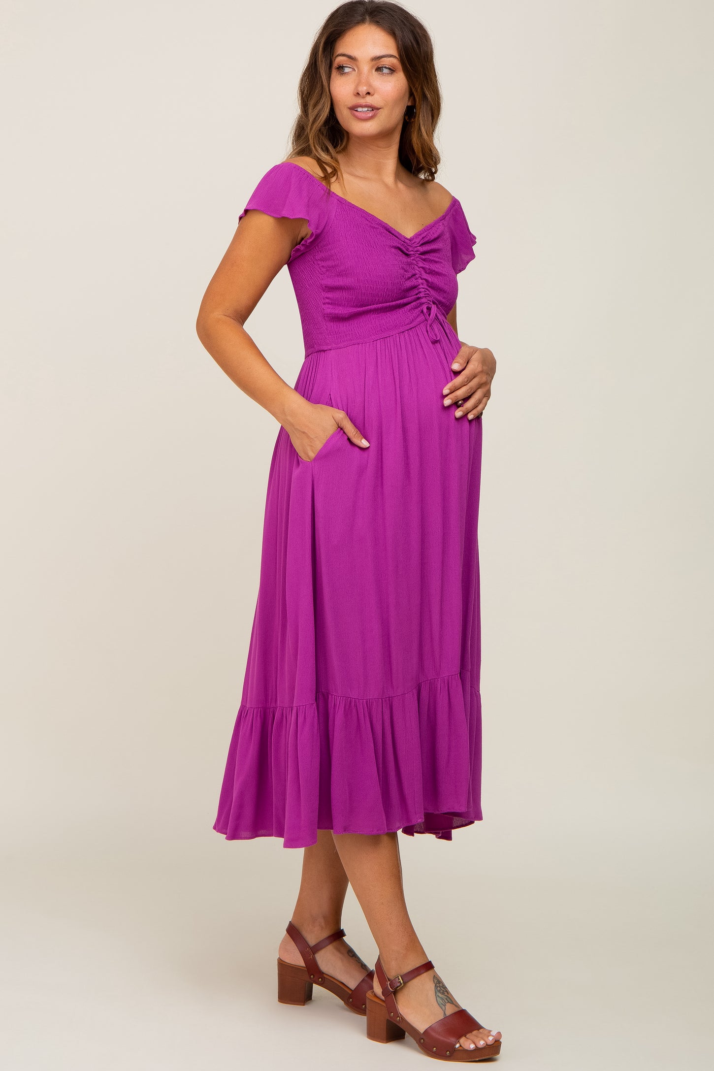 woman wearing a purple off-the-shoulder maternity dress with a ruched bodice and pockets, holding her baby bump