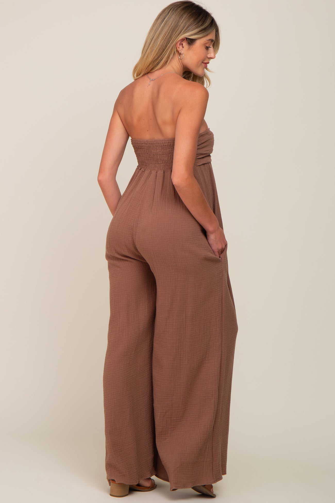 woman wearing a brown strapless smocked maternity jumpsuit with wide legs and pockets