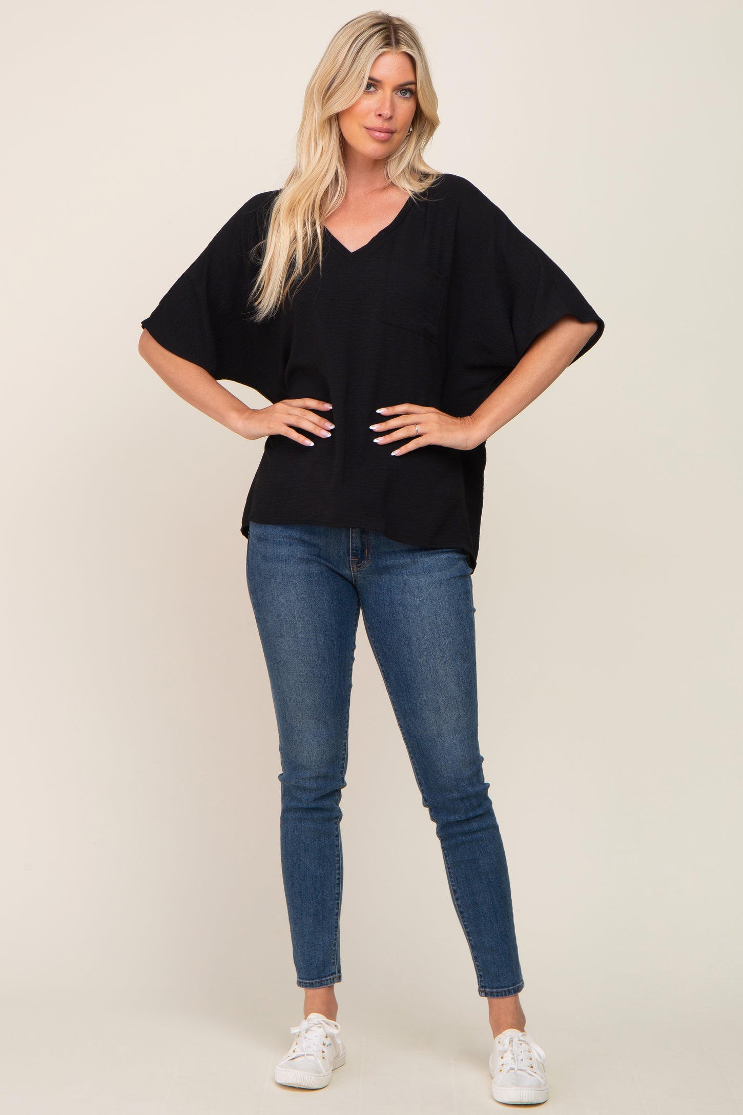 woman wearing a black loose fit v-neck top with short sleeves and a front pocket, paired with blue skinny jeans and white sneakers