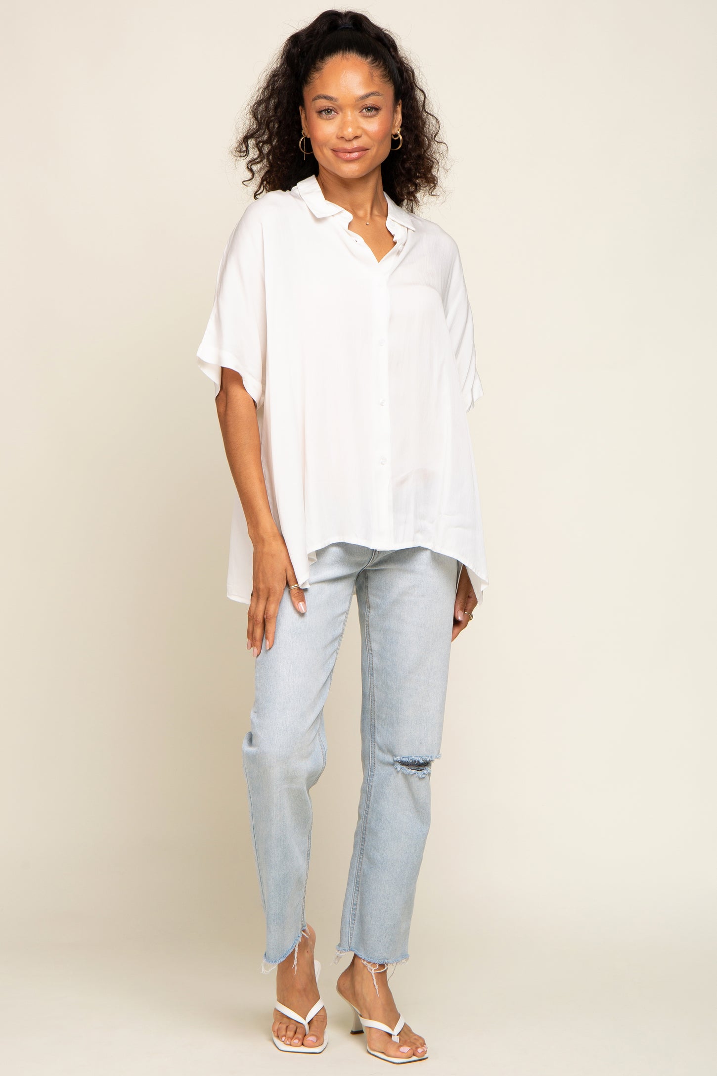 woman wearing a white oversized button-up shirt and light blue distressed jeans with white sandals.