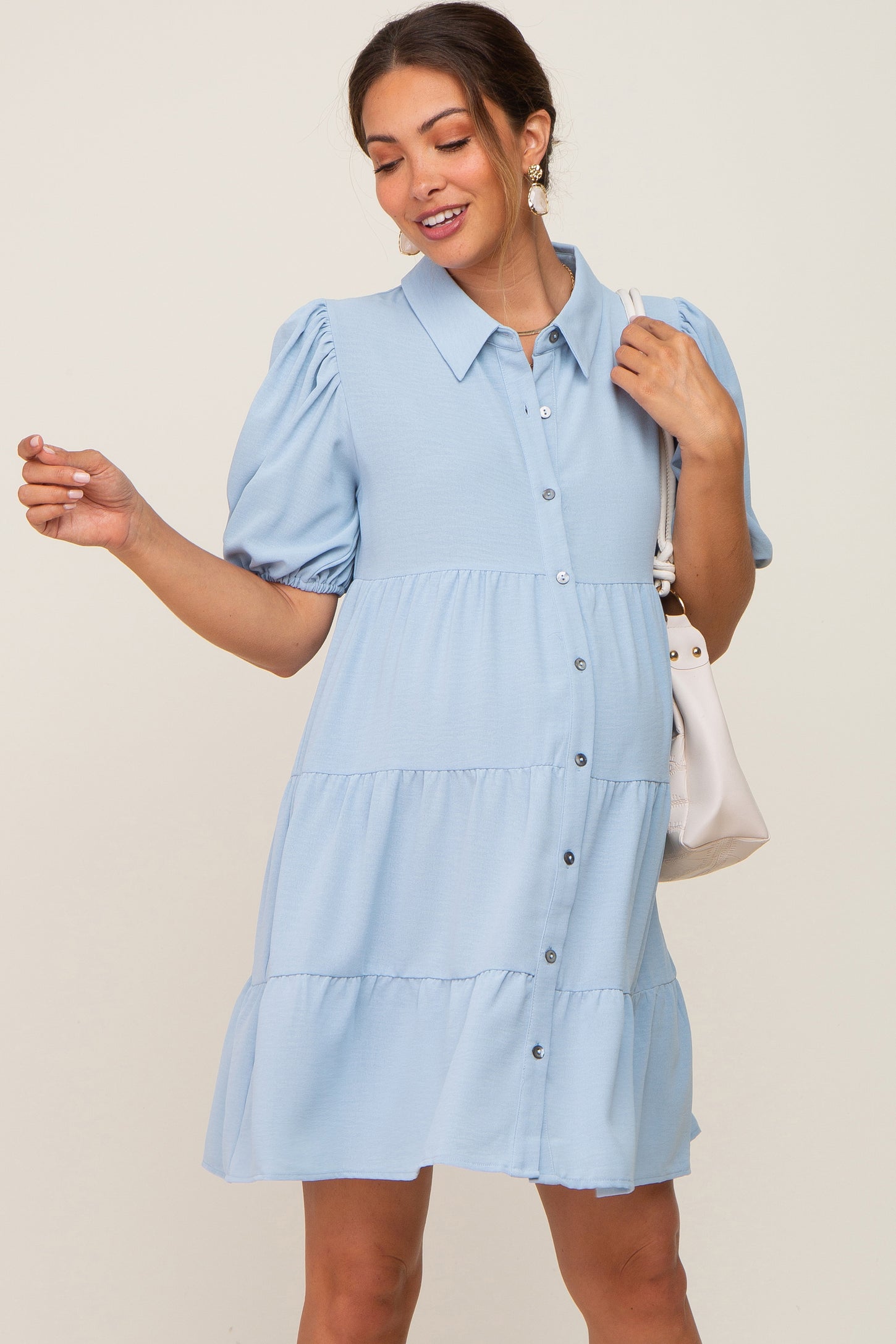 Woman wearing a light blue tiered maternity shirt dress with puff sleeves and a white shoulder bag