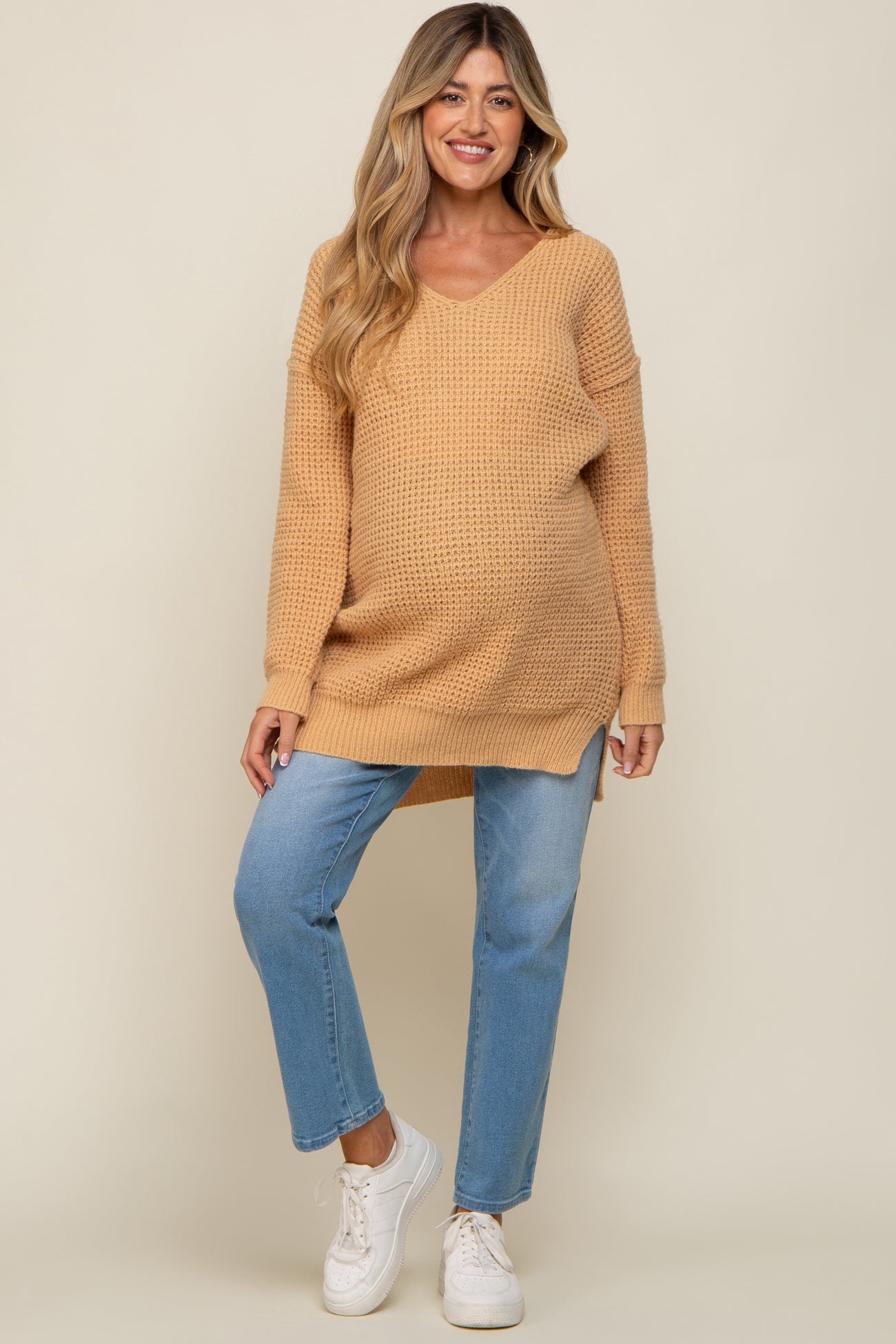 Pregnant woman wearing a tan waffle knit maternity sweater and blue jeans with white sneakers