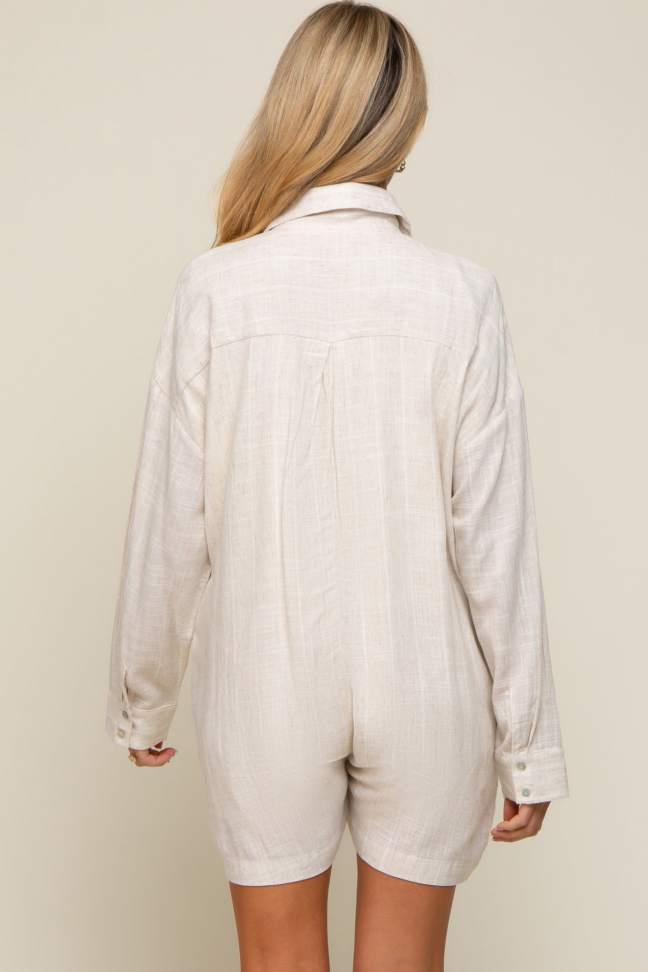 Back view of a woman wearing a beige linen long-sleeve romper with a collared neckline and button cuffs
