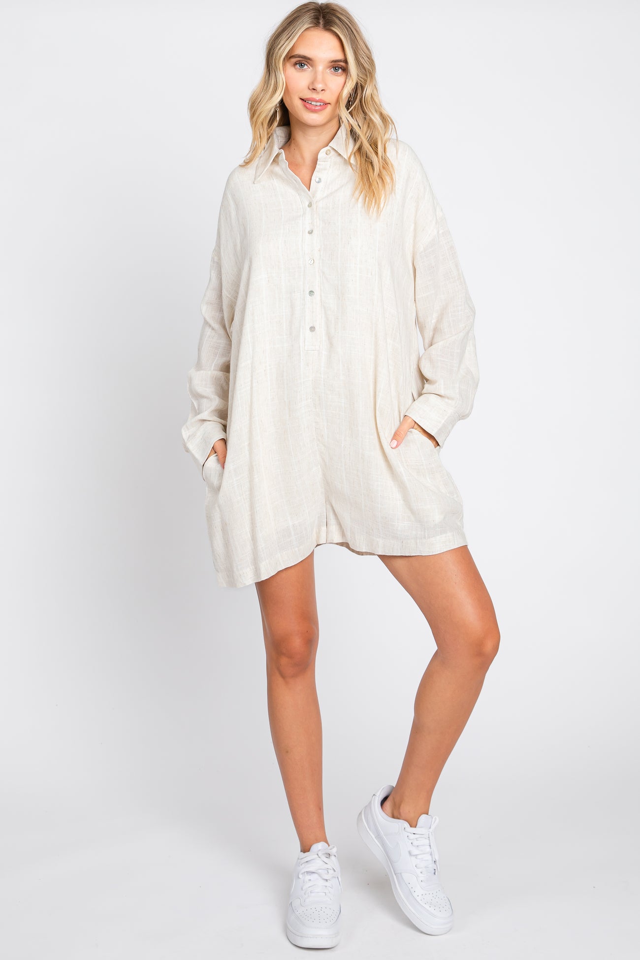 woman wearing a cream linen long sleeve romper with buttons and pockets paired with white sneakers