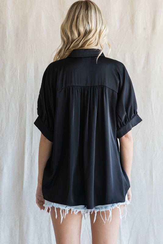 Back view of a woman wearing a black short sleeve satin button down shirt with pleated detail and denim shorts