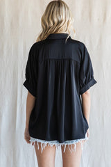 Back view of a woman wearing a black short sleeve satin button down shirt with pleated detail and denim shorts