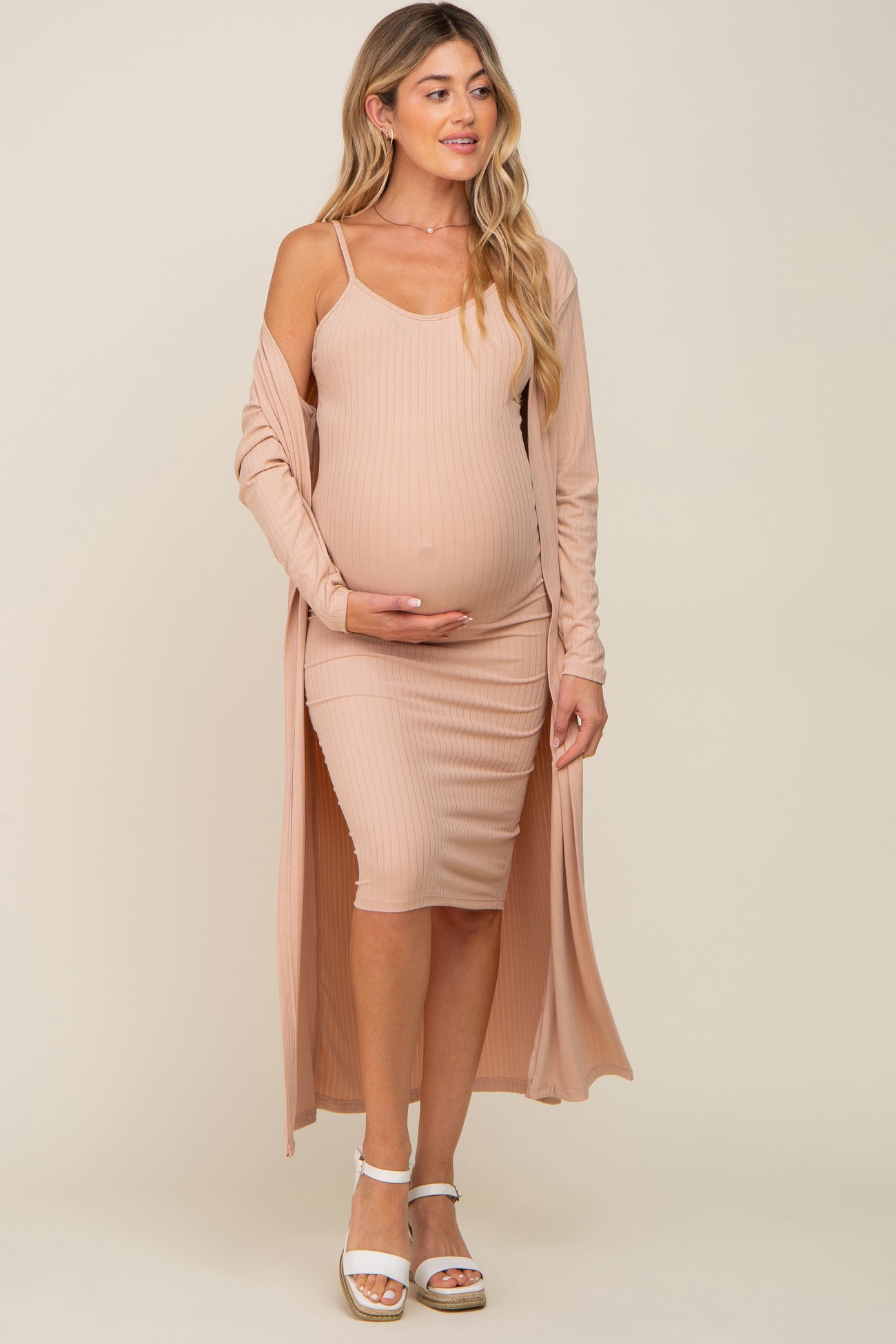 woman wearing a light pink maternity dress with ribbed texture and a matching long cardigan, cradling her baby bump