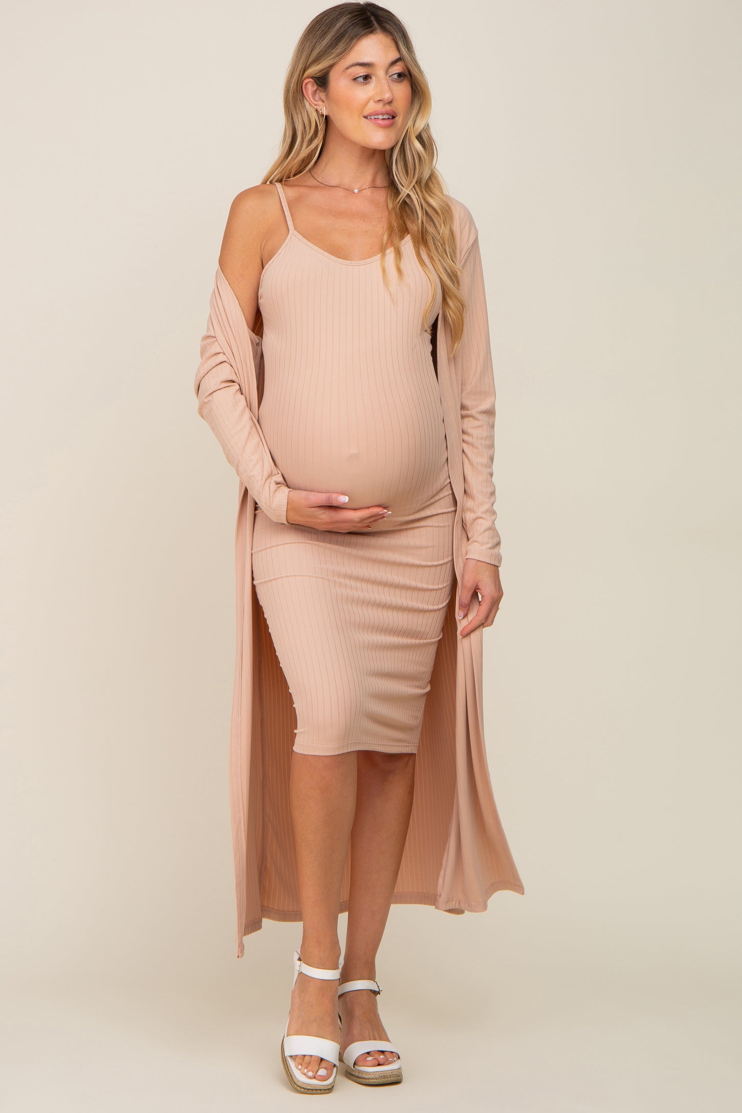 woman wearing a light pink maternity dress with ribbed texture and a matching long cardigan, cradling her baby bump