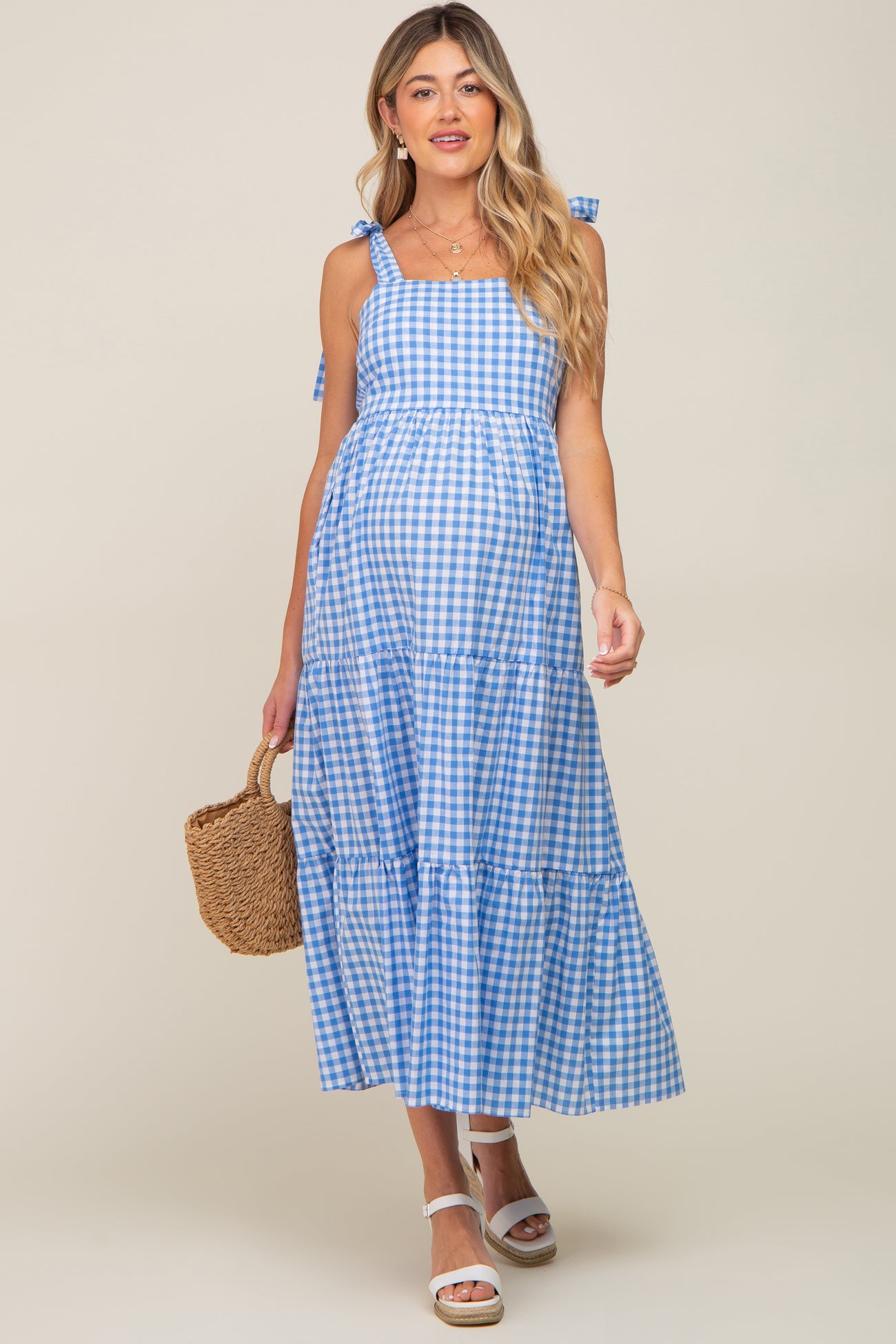 woman wearing a blue and white gingham maternity maxi dress with tied straps and holding a woven bag, showing her baby bump