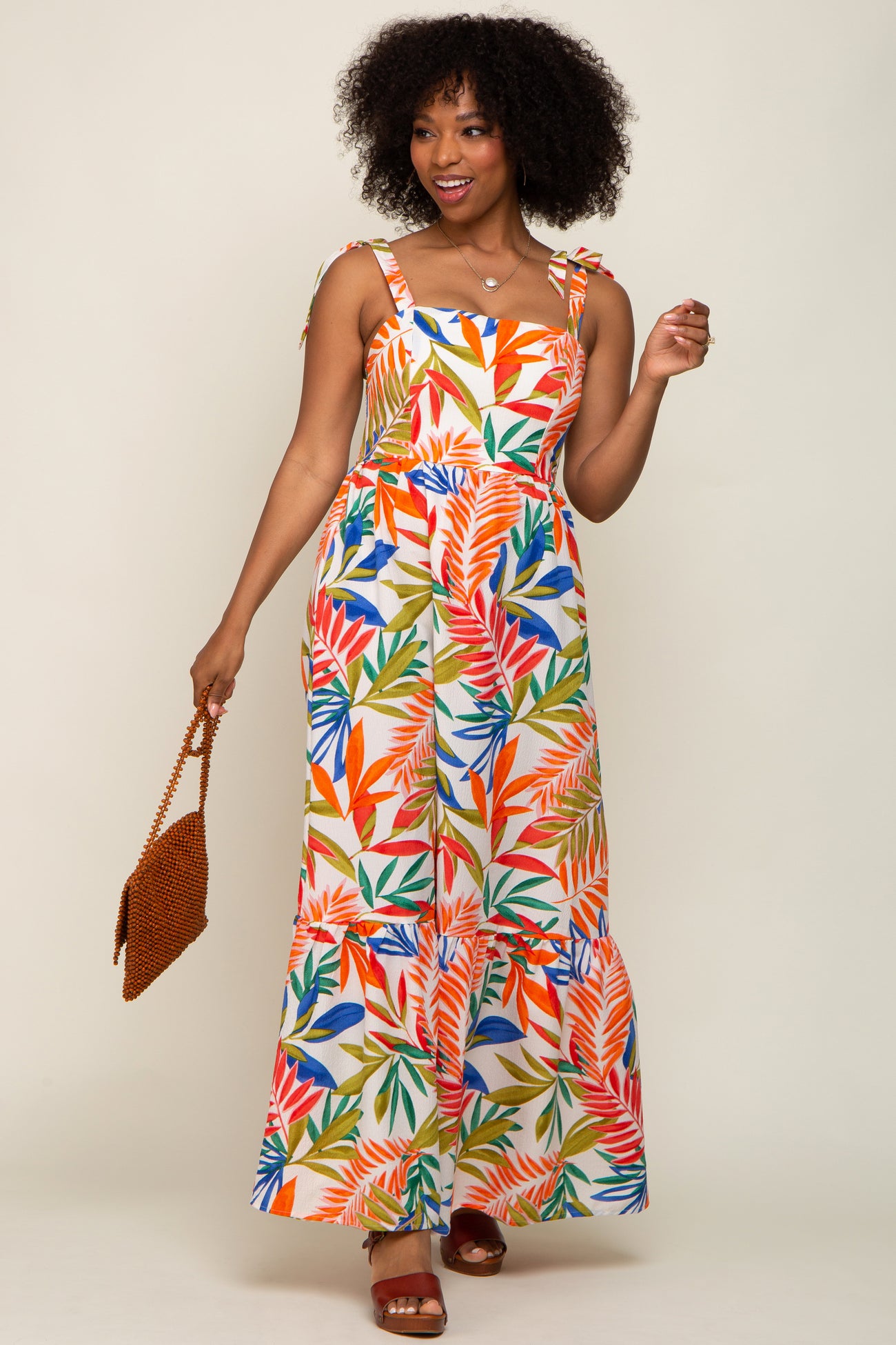 woman wearing a tropical leaf print maxi dress with tie straps and holding a brown beaded handbag