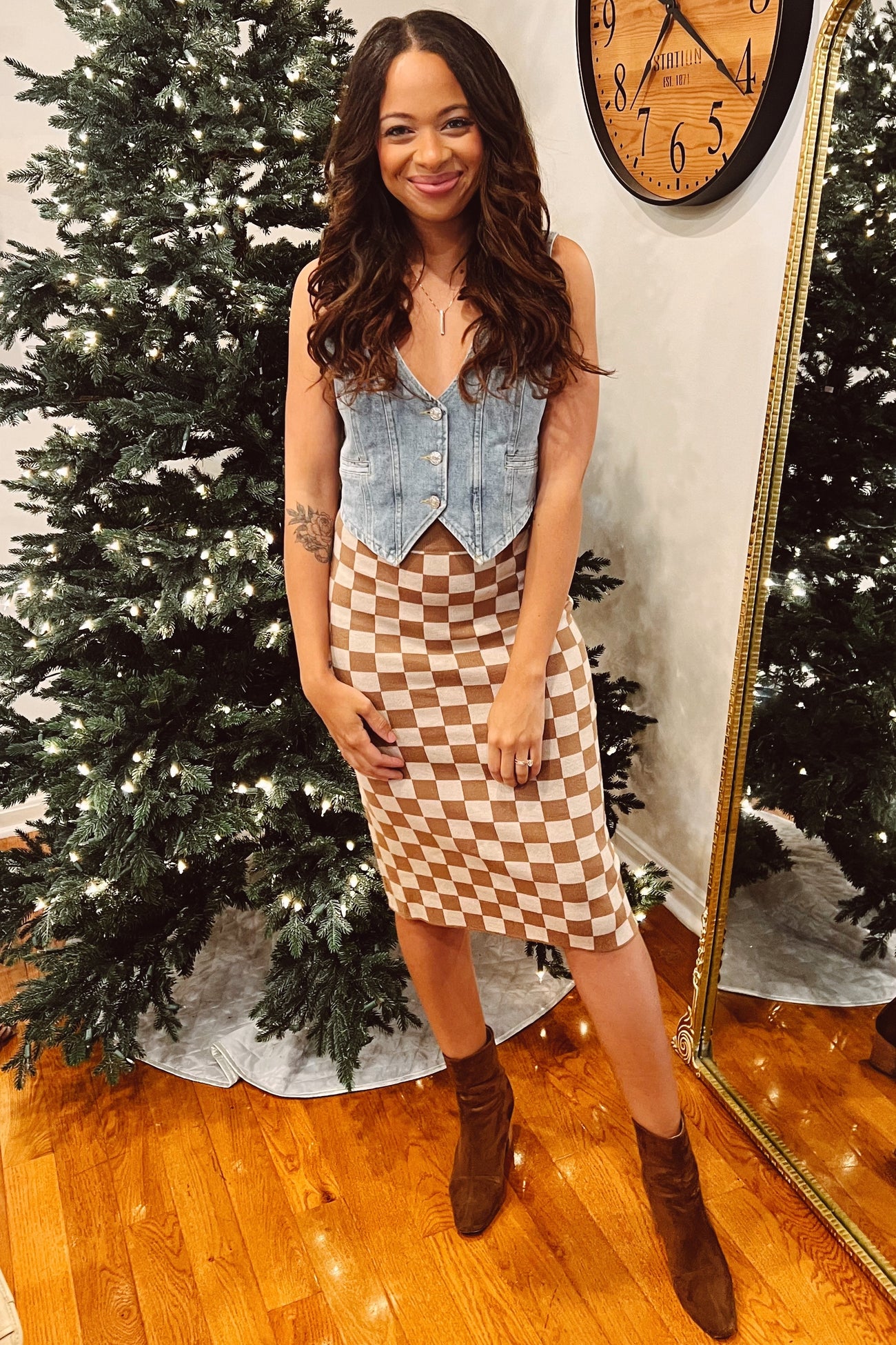 woman wearing a denim maternity vest and a beige checkered midi skirt, posing in front of a festive background