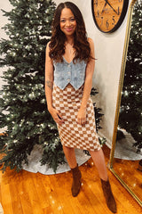 woman wearing a denim maternity vest and a beige checkered midi skirt, posing in front of a festive background