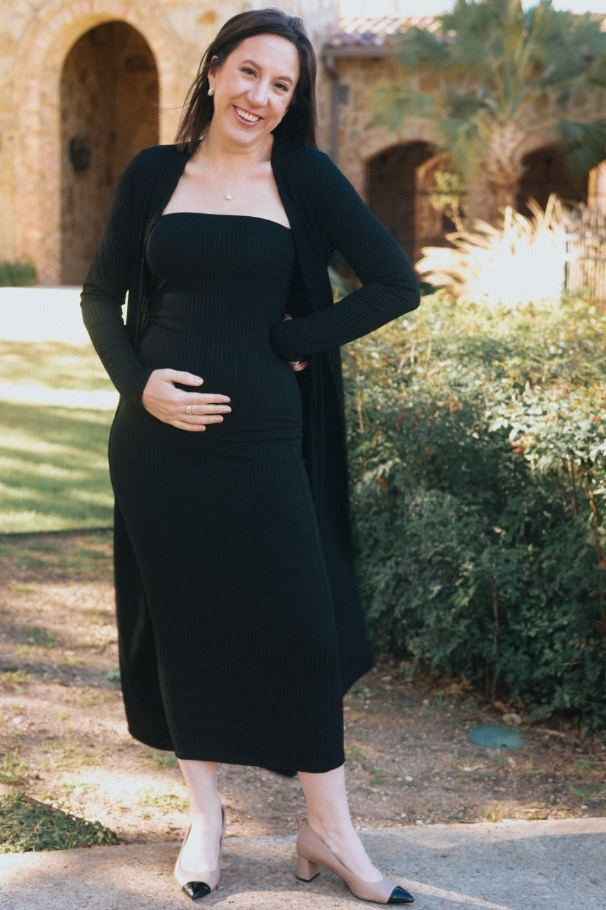 woman wearing a black ribbed maternity dress and long cardigan, holding her baby bump outdoors