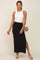 woman wearing a white ribbed sleeveless top and a black maxi skirt with a side slit, holding a small handbag