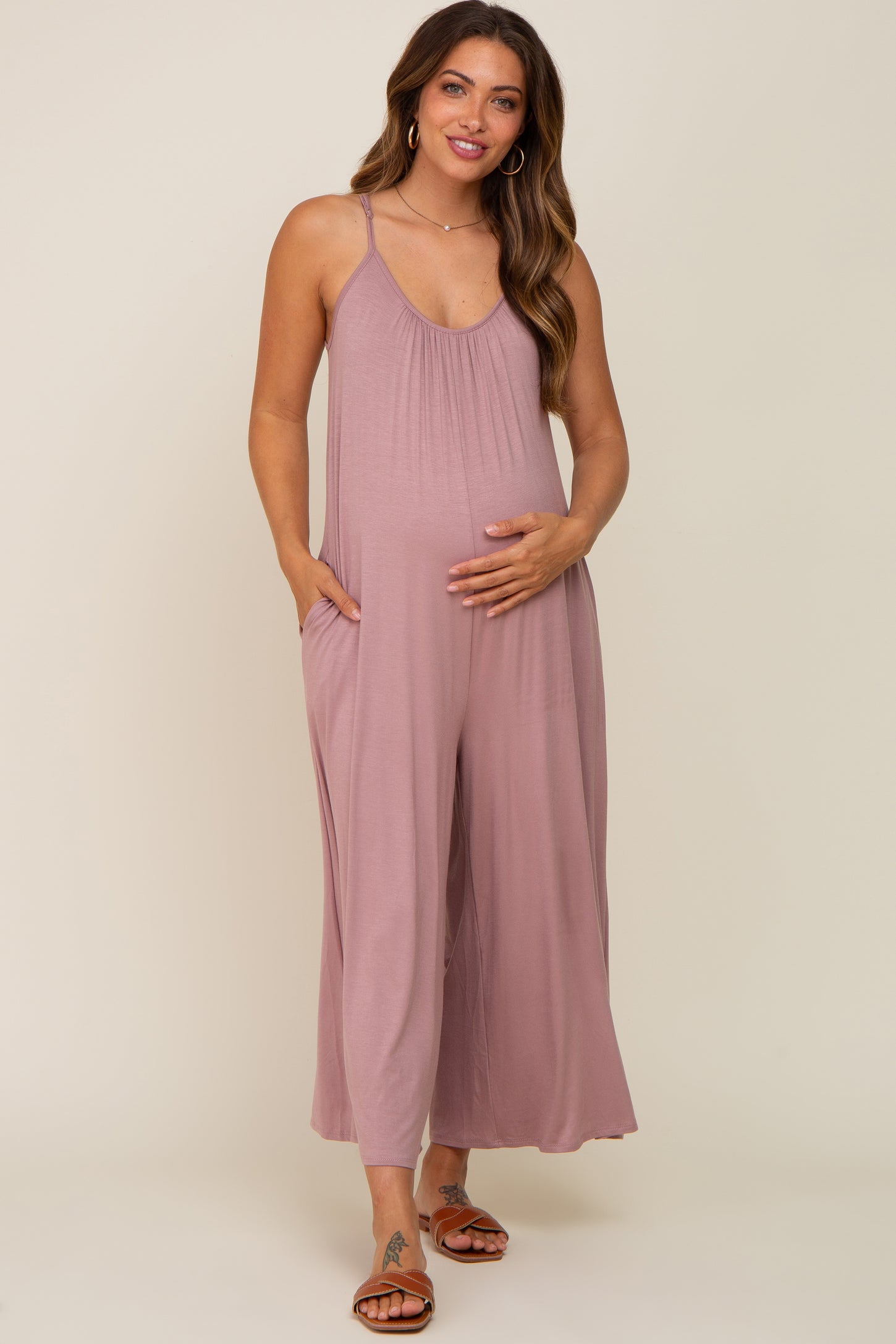 woman wearing a mauve sleeveless maternity jumpsuit with pockets and a visible baby bump, standing and smiling