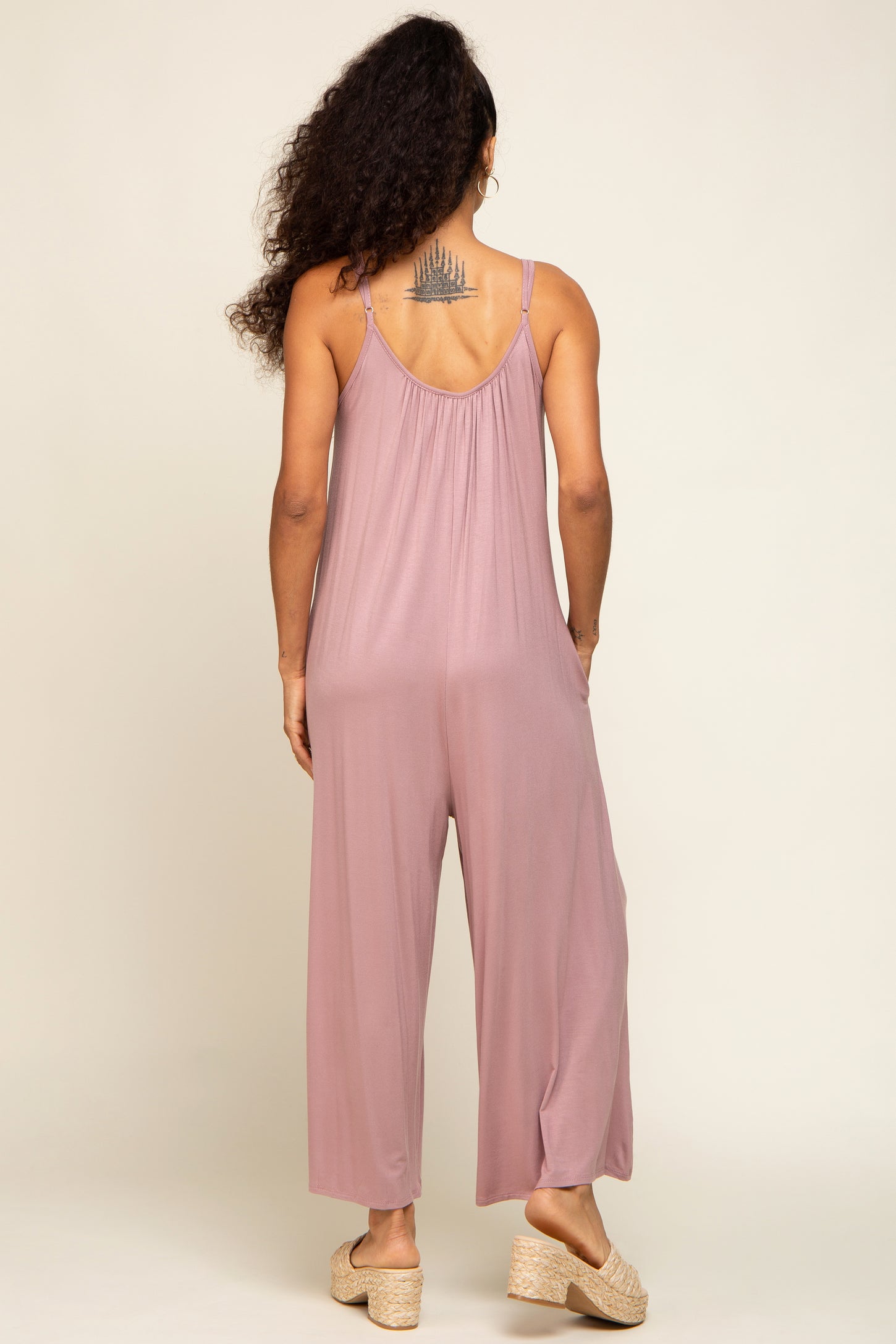 woman wearing a mauve sleeveless jumpsuit with loose fitting legs and a strappy back, standing with her back to the camera