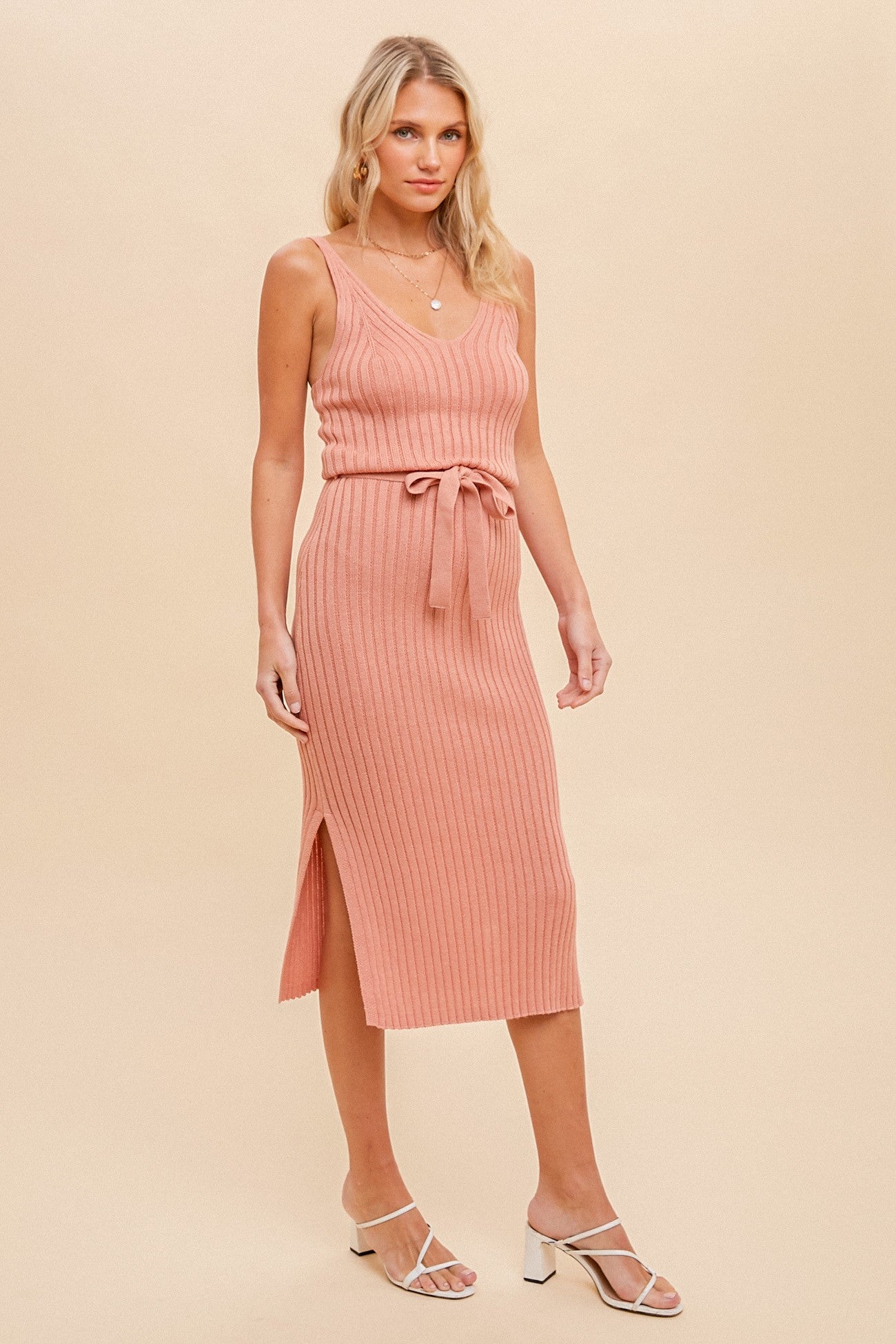 woman wearing a salmon pink ribbed midi dress with a tie belt and side slit, paired with white strappy sandals