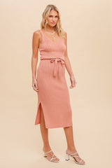 woman wearing a salmon pink ribbed midi dress with a tie belt and side slit, paired with white strappy sandals