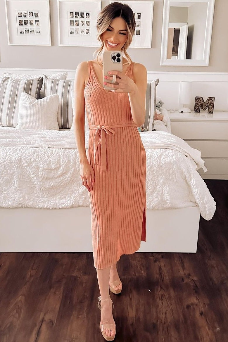 woman wearing a coral ribbed midi dress with a tie waist and side slit, standing in a stylish bedroom setting