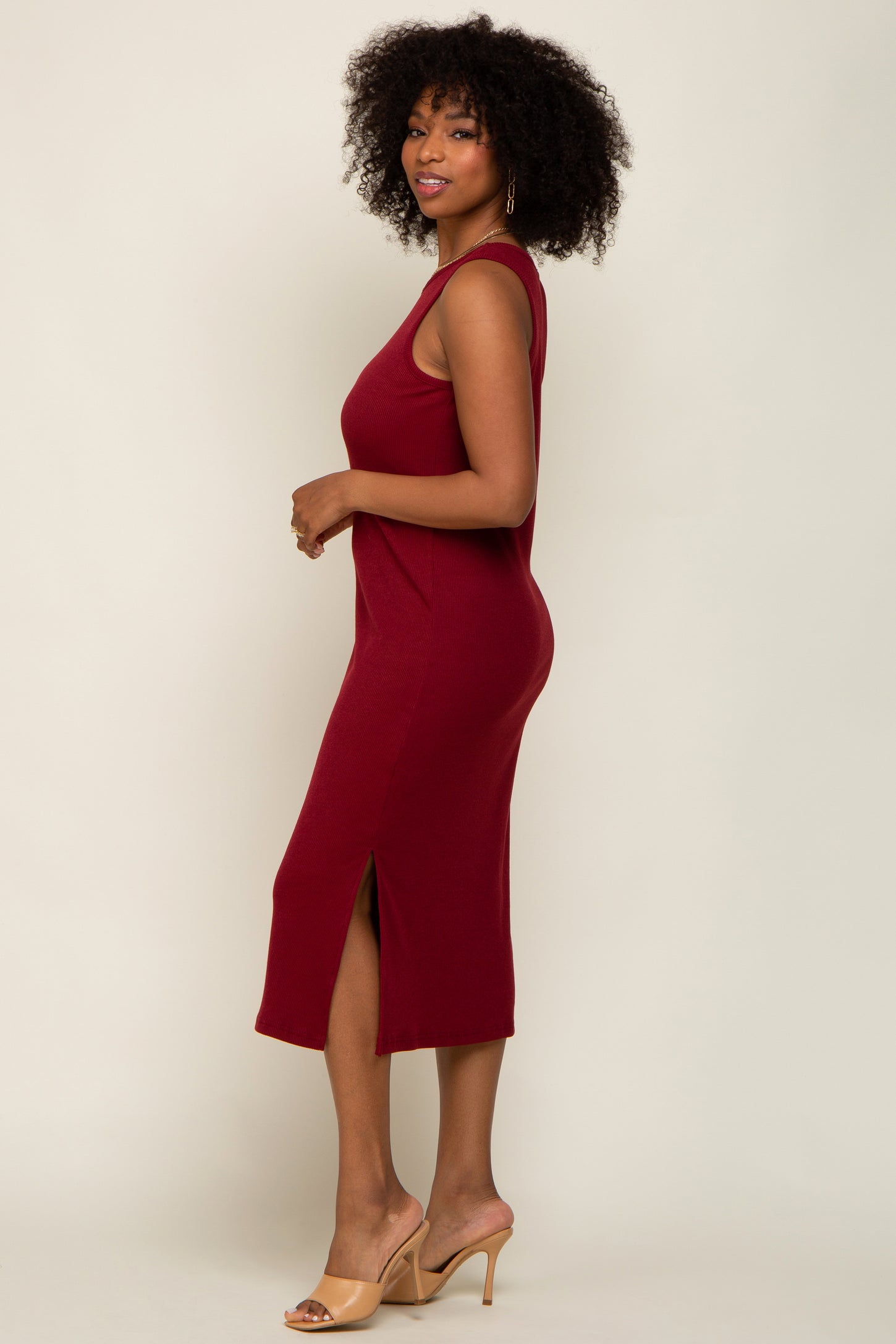 woman with curly hair wearing a sleeveless burgundy midi dress with a side slit and tan heels