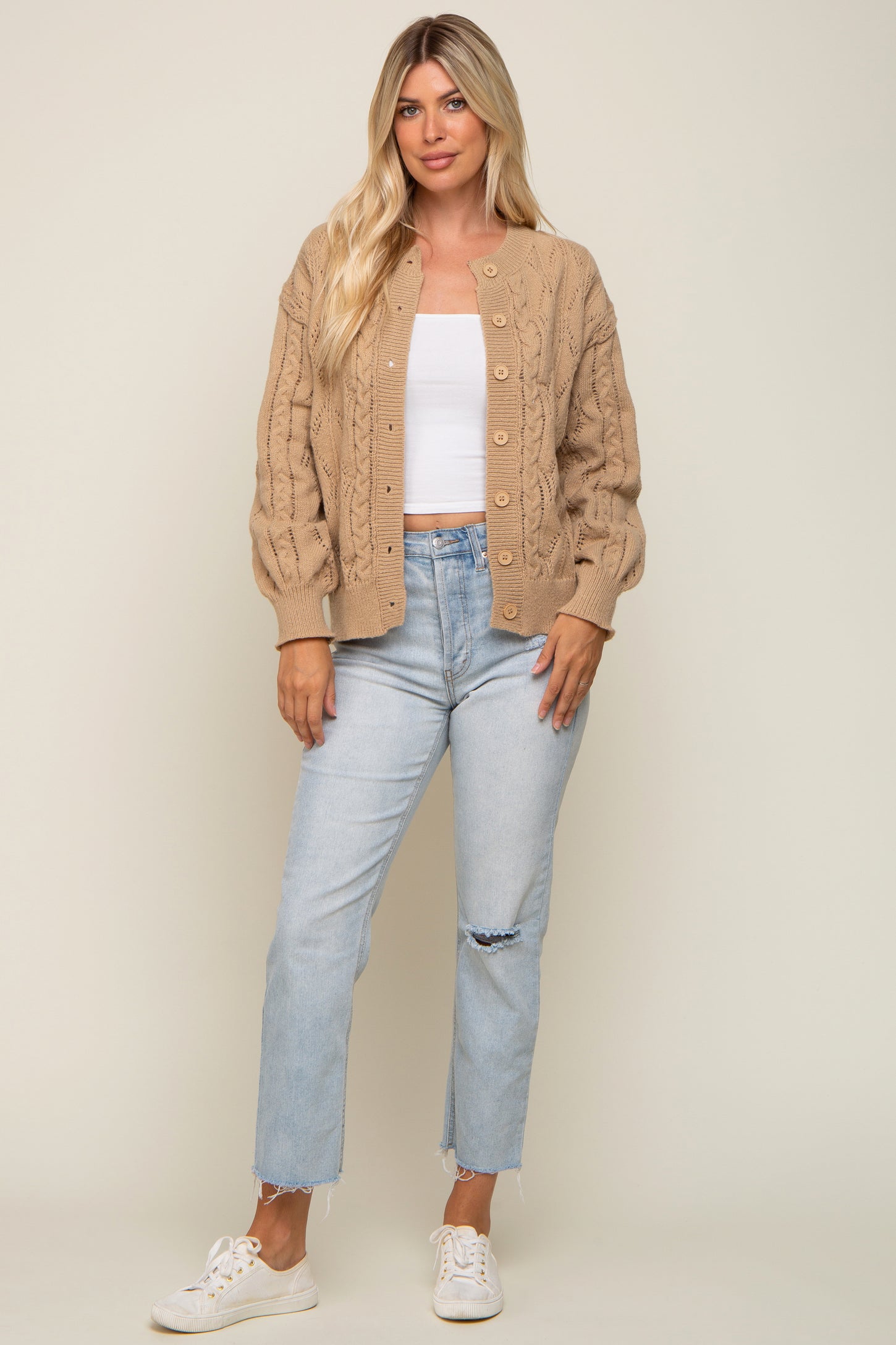 Woman wearing a tan cable knit cardigan, white crop top, and light wash distressed jeans