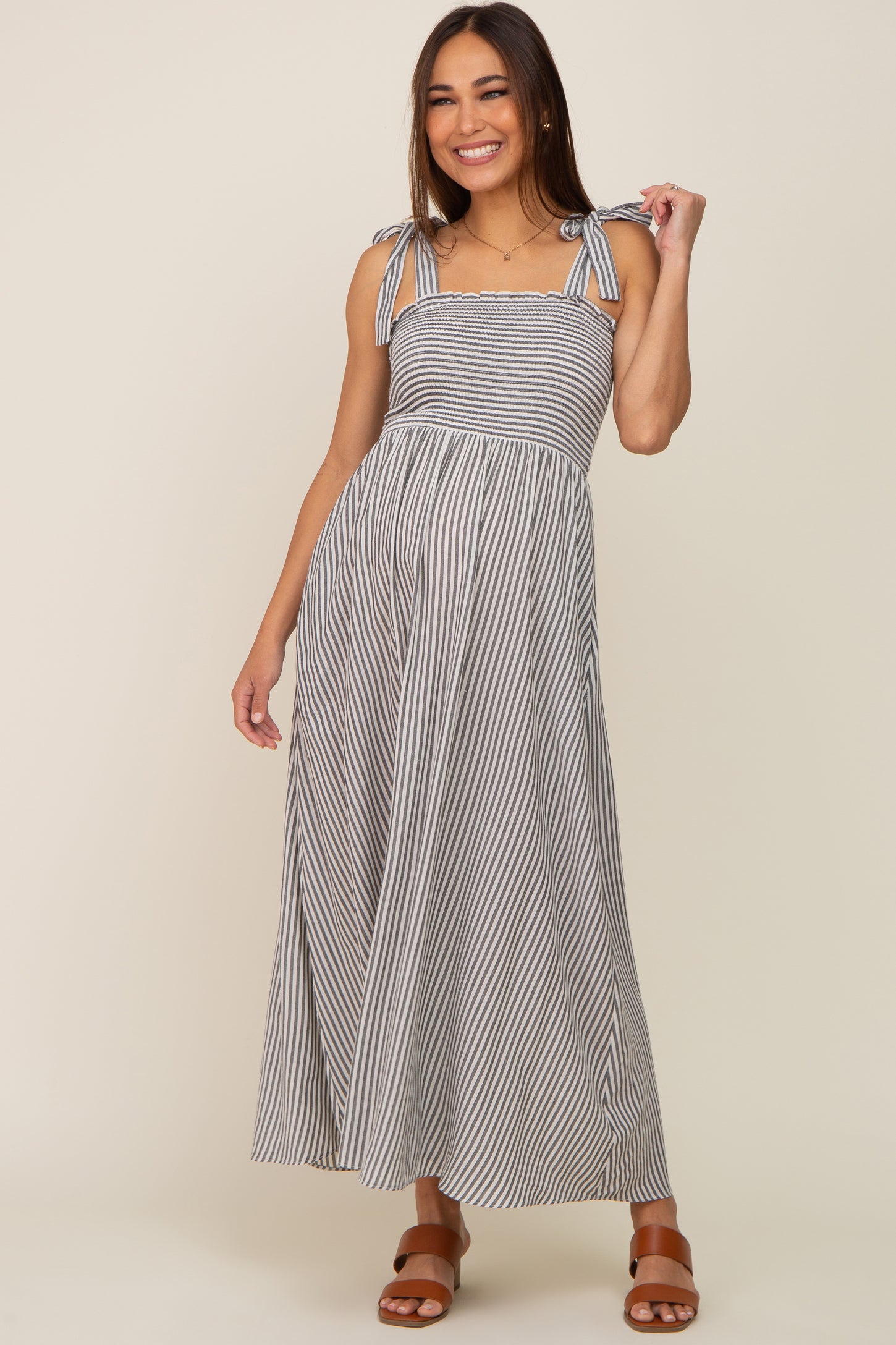 smiling woman wearing a grey and white striped smocked maternity maxi dress with tie straps and brown sandals