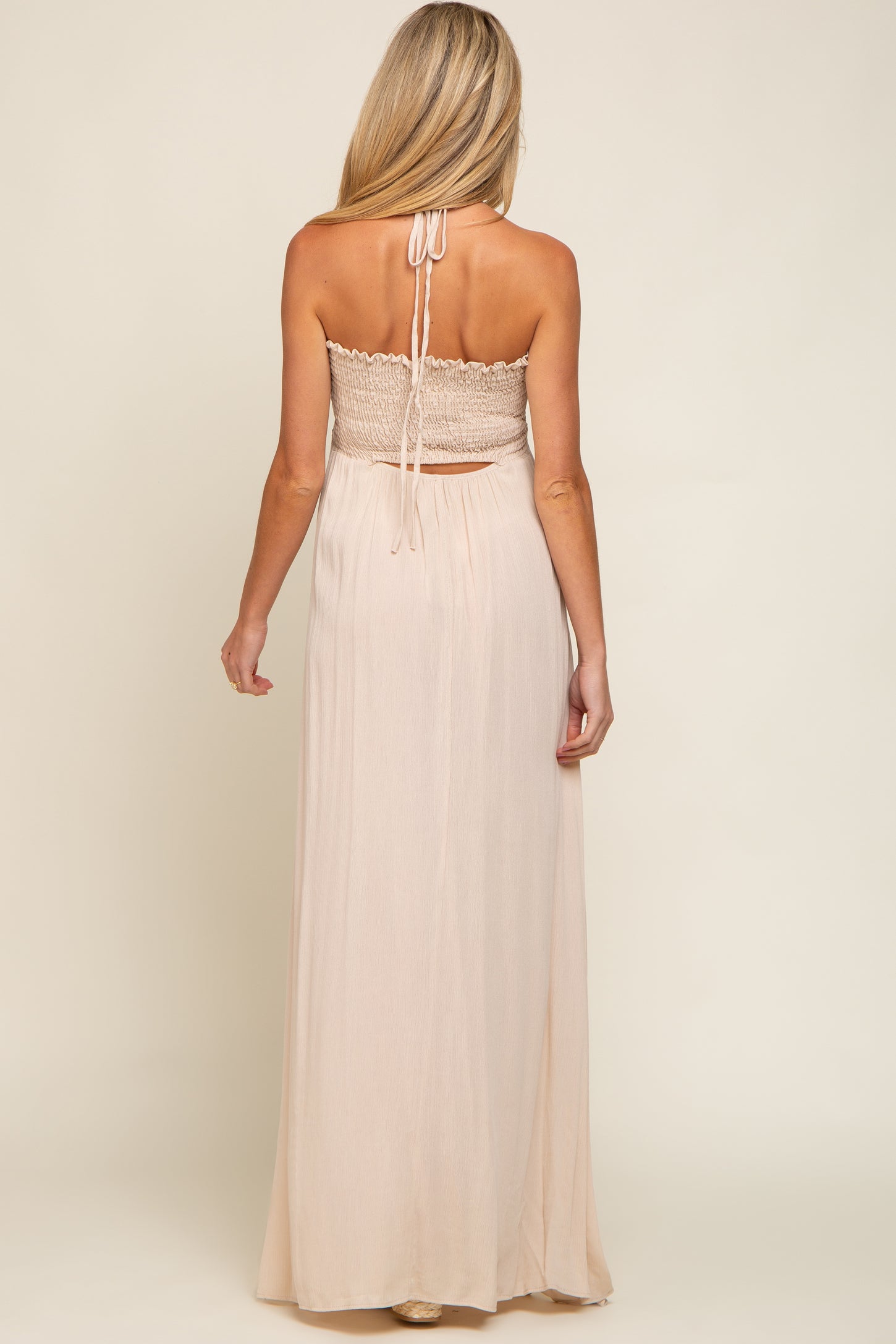 woman wearing a light peach maternity maxi dress with a smocked bodice and halter neckline, showing off her baby bump rear view