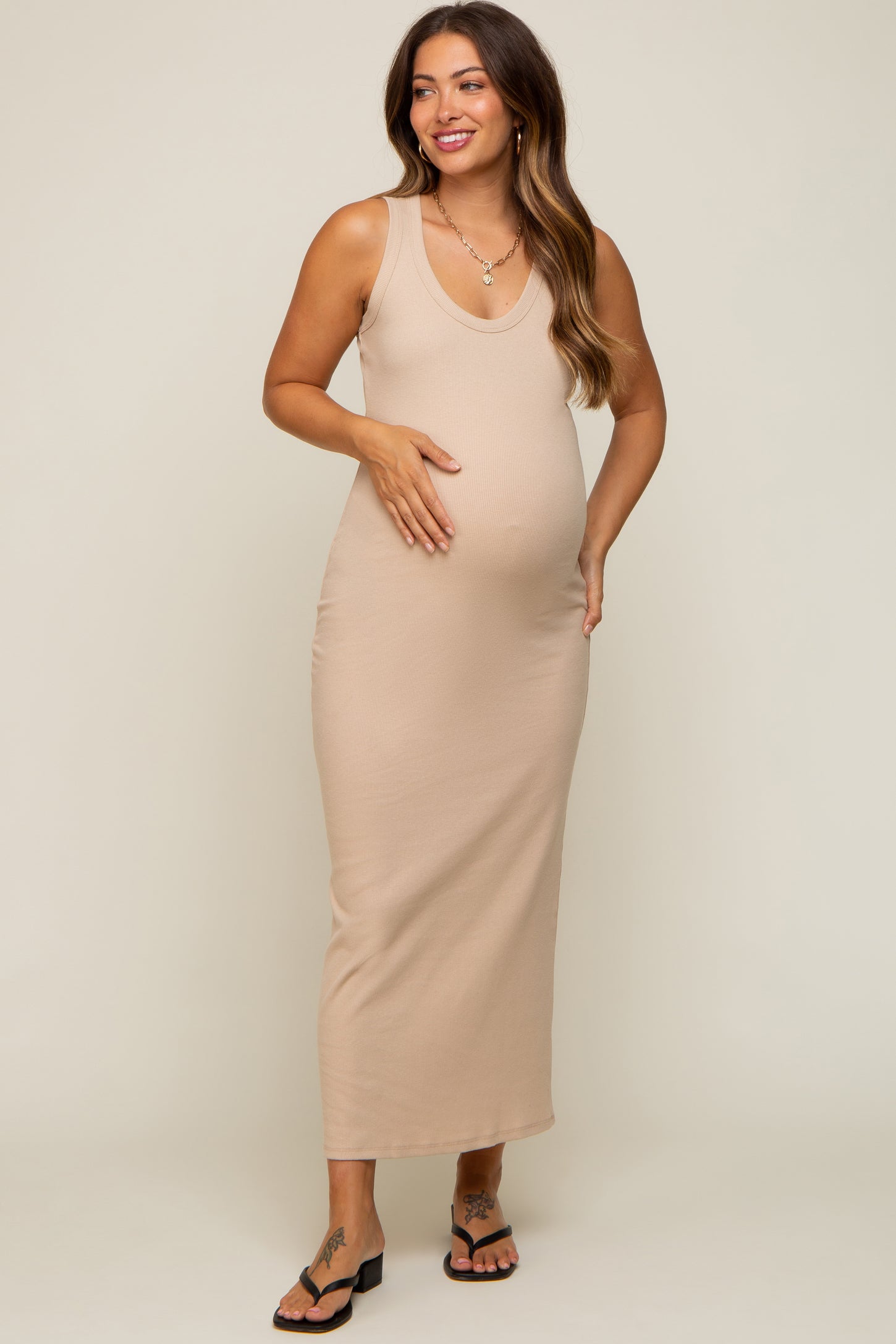 woman wearing a beige sleeveless maternity dress and holding her baby bump, styled with black sandals