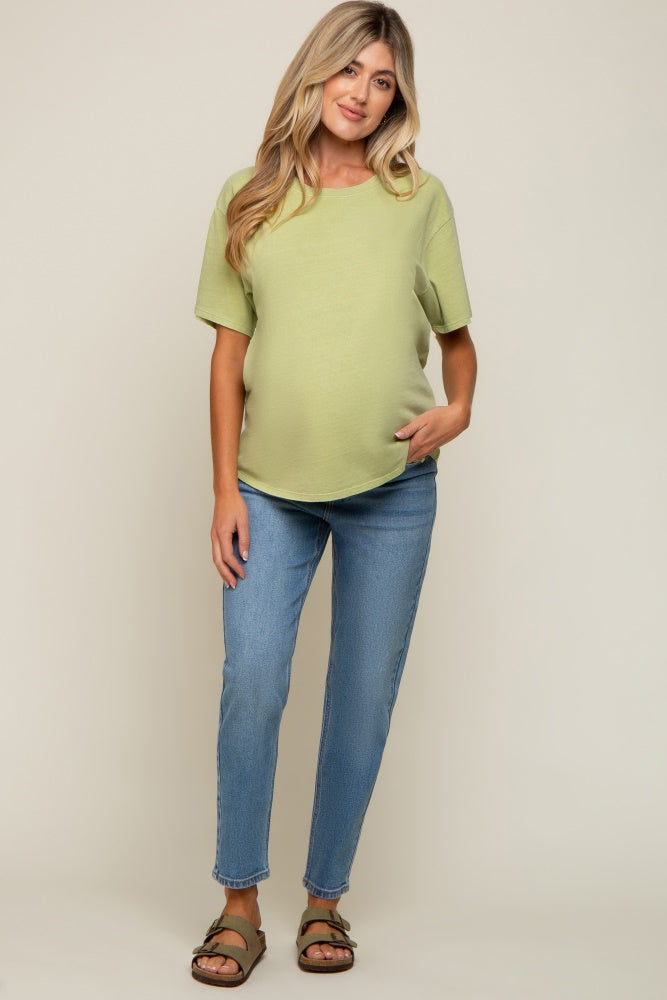 woman wearing a light green maternity top and blue jeans, showcasing her baby bump, standing with one hand in her pocket