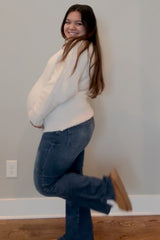 smiling pregnant woman in white sweater and blue jeans posing against a neutral wall