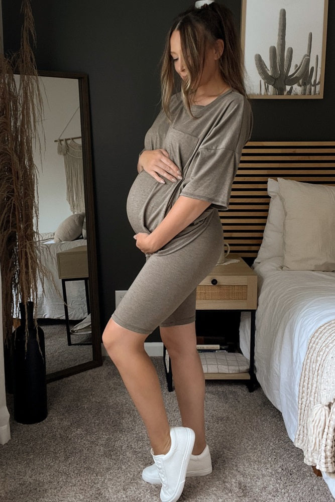 woman wearing a taupe maternity t-shirt dress and holding her baby bump while standing in a bedroom setting