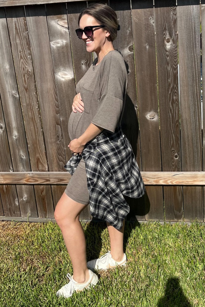 woman wearing a brown maternity dress with a tied black and white plaid shirt and holding her baby bump