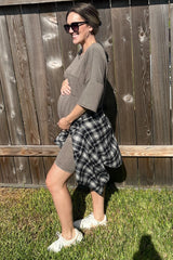 woman wearing a brown maternity dress with a tied black and white plaid shirt and holding her baby bump