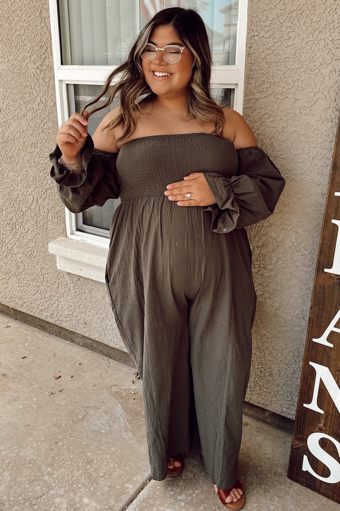 woman wearing an olive green off-the-shoulder maternity jumpsuit, smiling and holding her baby bump