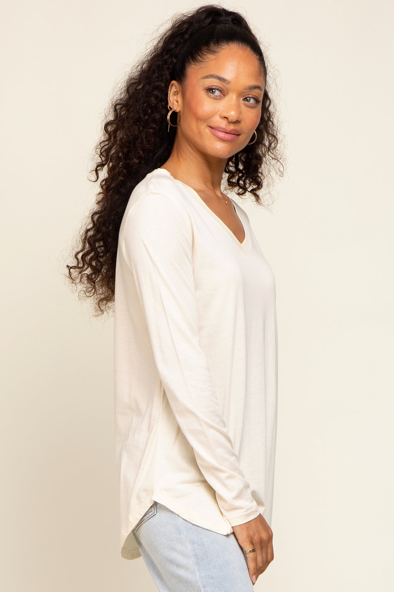 Woman wearing a cream long-sleeve v-neck shirt and light wash jeans against a neutral background