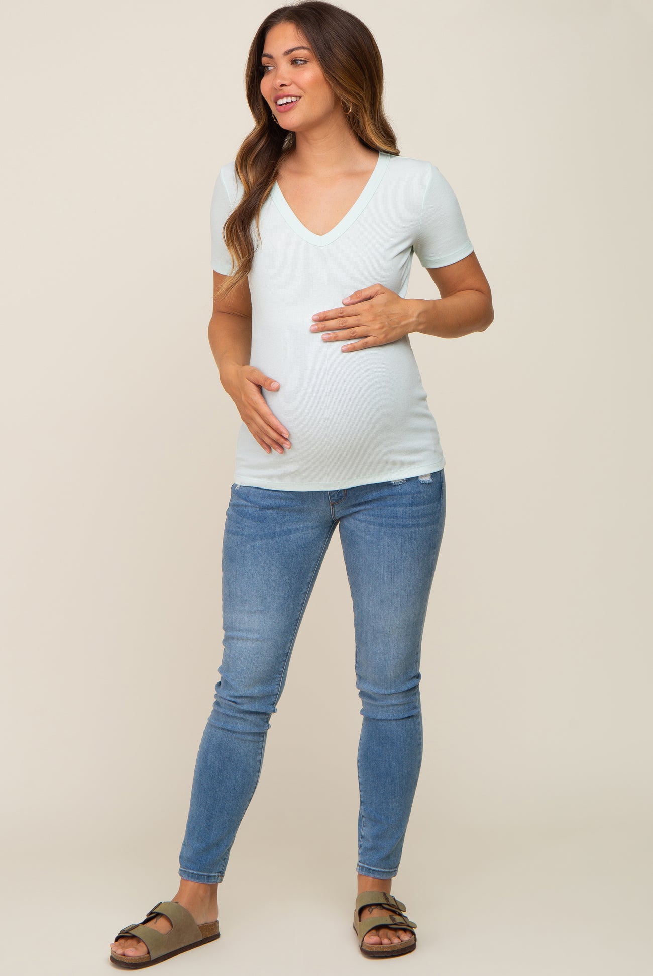 woman wearing a light green v-neck maternity shirt and blue jeans, cradling her baby bump