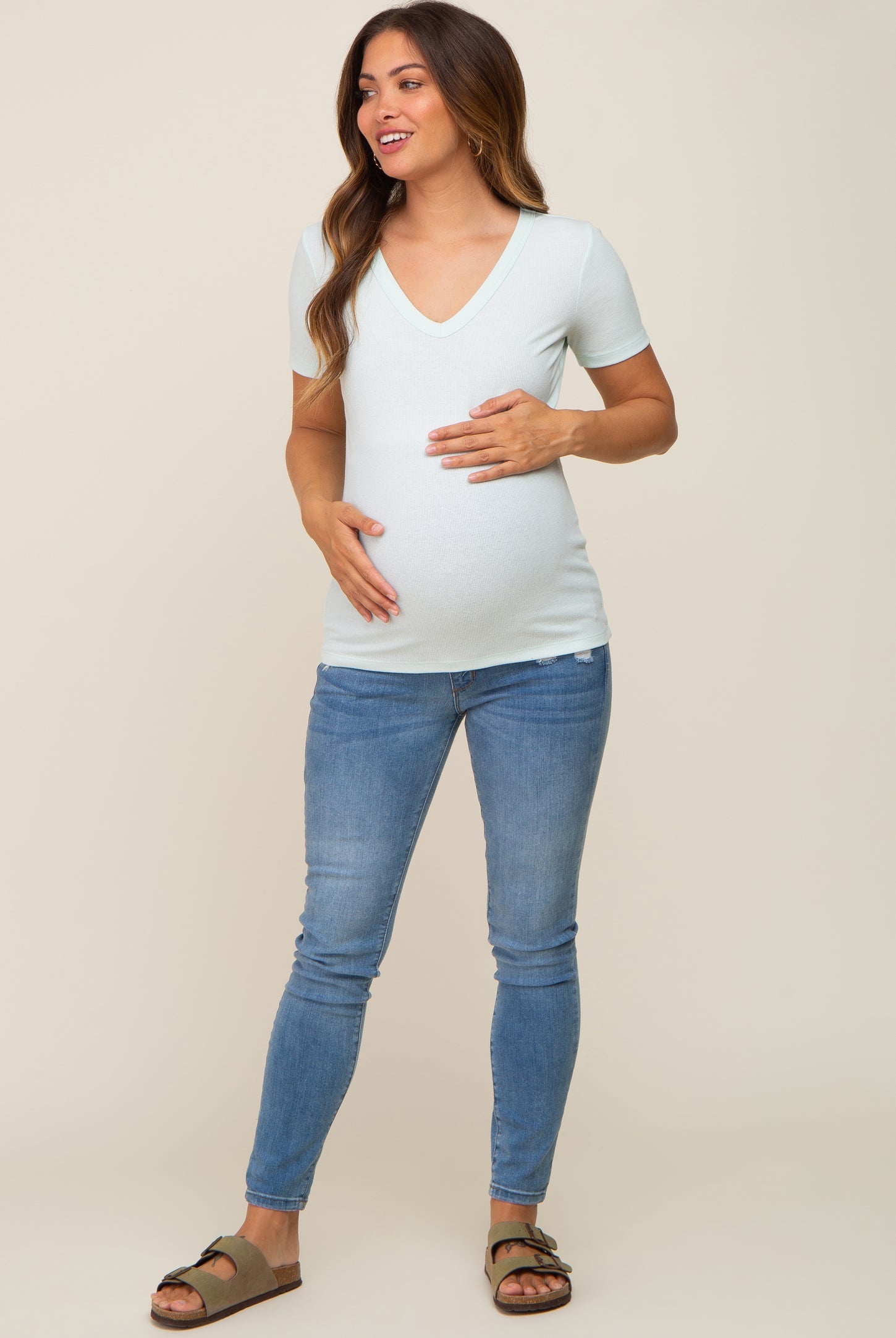woman wearing a light green v-neck maternity shirt and blue jeans, cradling her baby bump